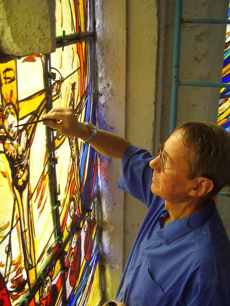 Un homme en chemise bleue travaille sur un vitrail, à l'aide d'un pinceau, à l'intérieur d'un bâtiment.