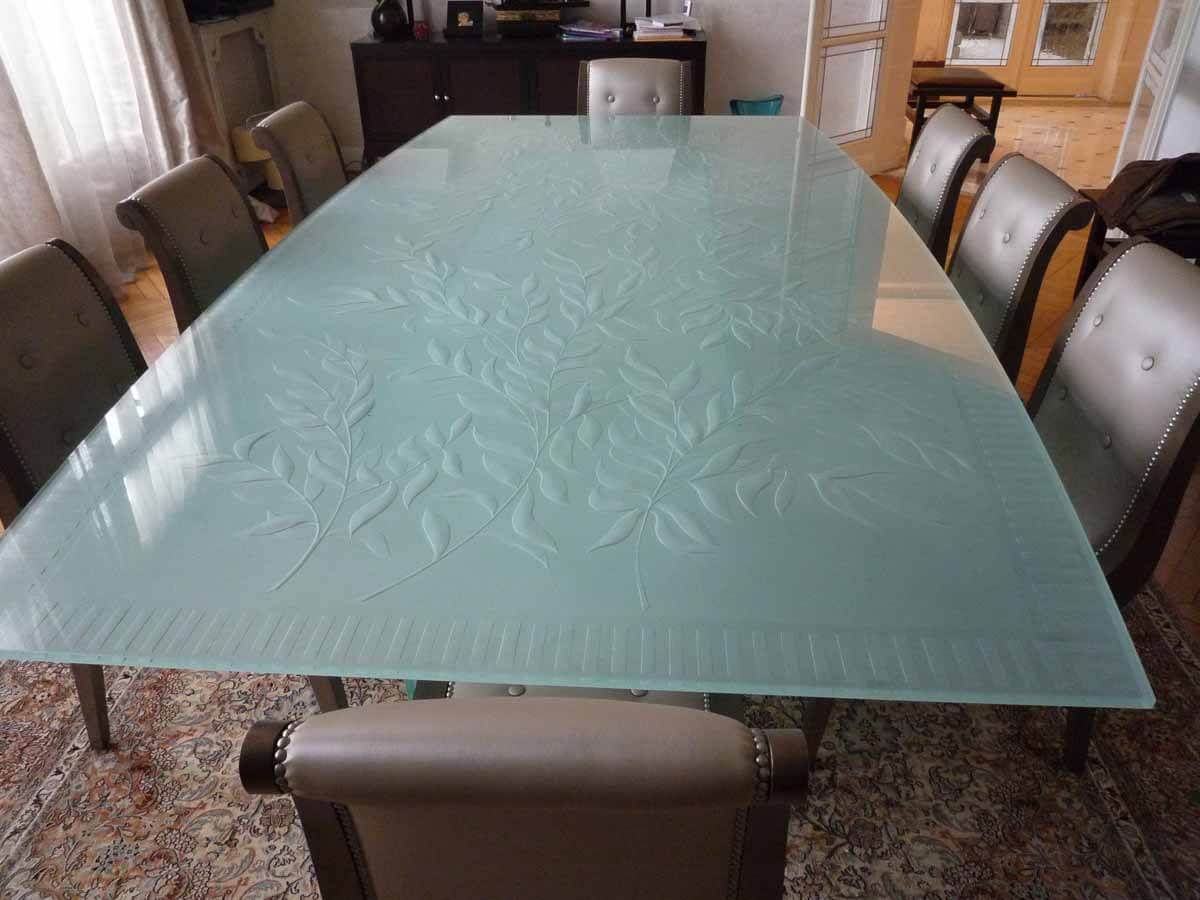 Table de salle à manger en verre ornée de motifs floraux gravés, entourée de chaises en cuir gris.