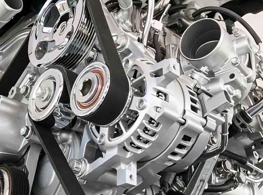 Imagem em close-up de um motor de carro com correias, polias e alternador, predominantemente prateado com correias pretas.