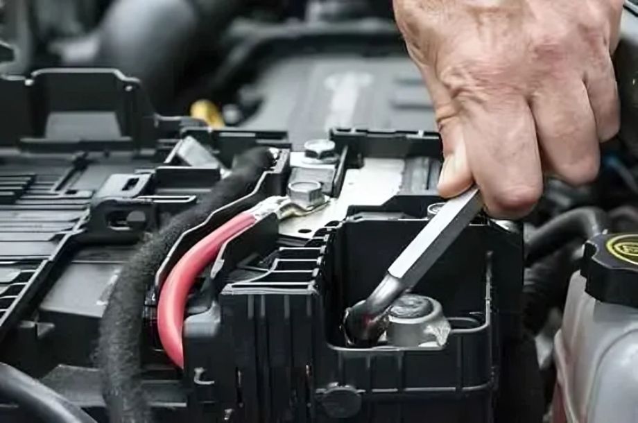 Mão usando uma chave inglesa para soltar um parafuso no terminal da bateria de um carro no compartimento do motor.