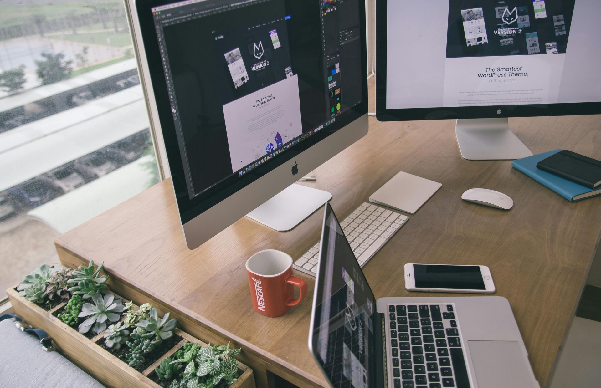 A desk with two computer monitors, a laptop, a red mug, a phone, and a long wooden planter filled with succulents.