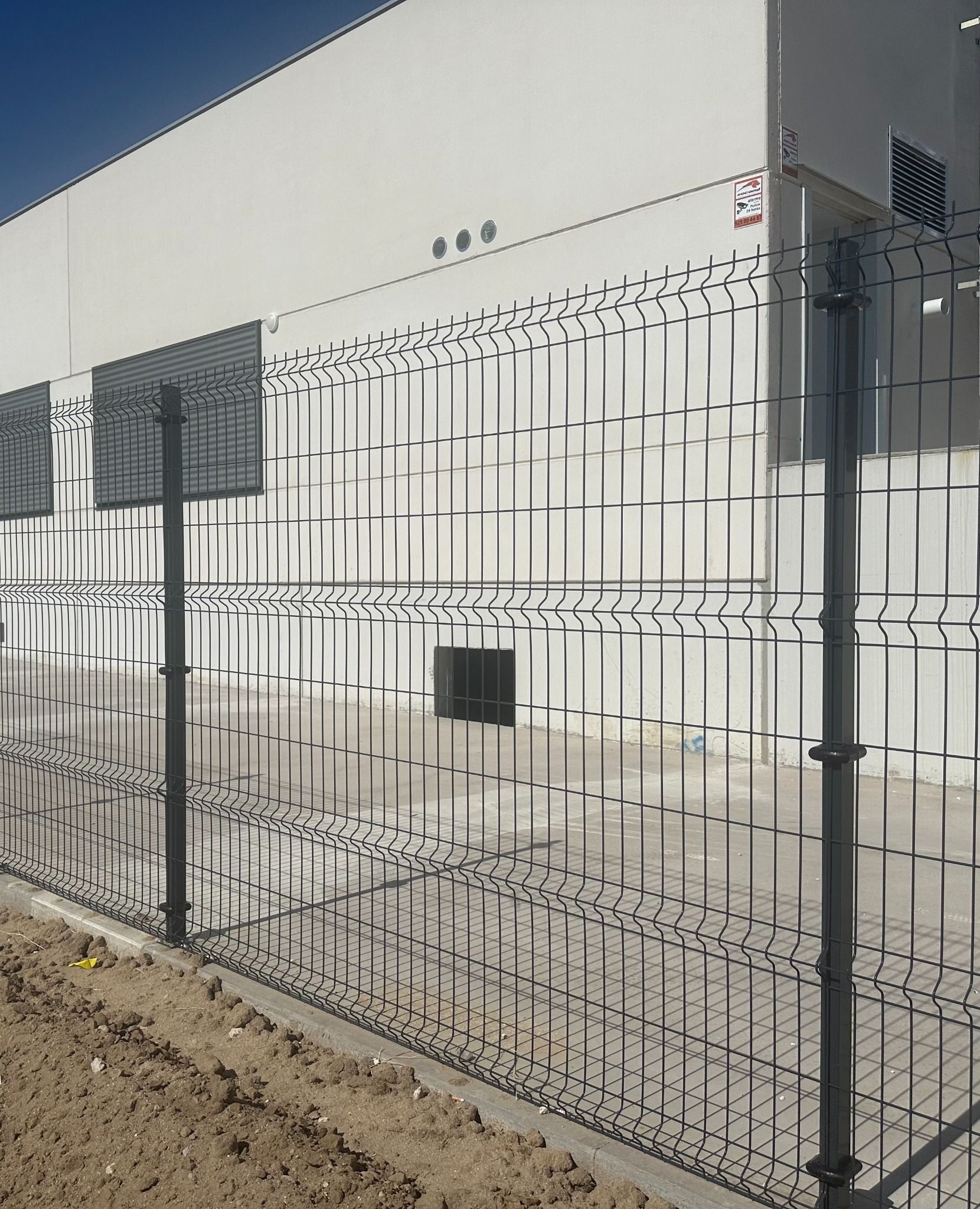 Valla de seguridad de metal gris frente a un edificio industrial blanco en un día soleado.