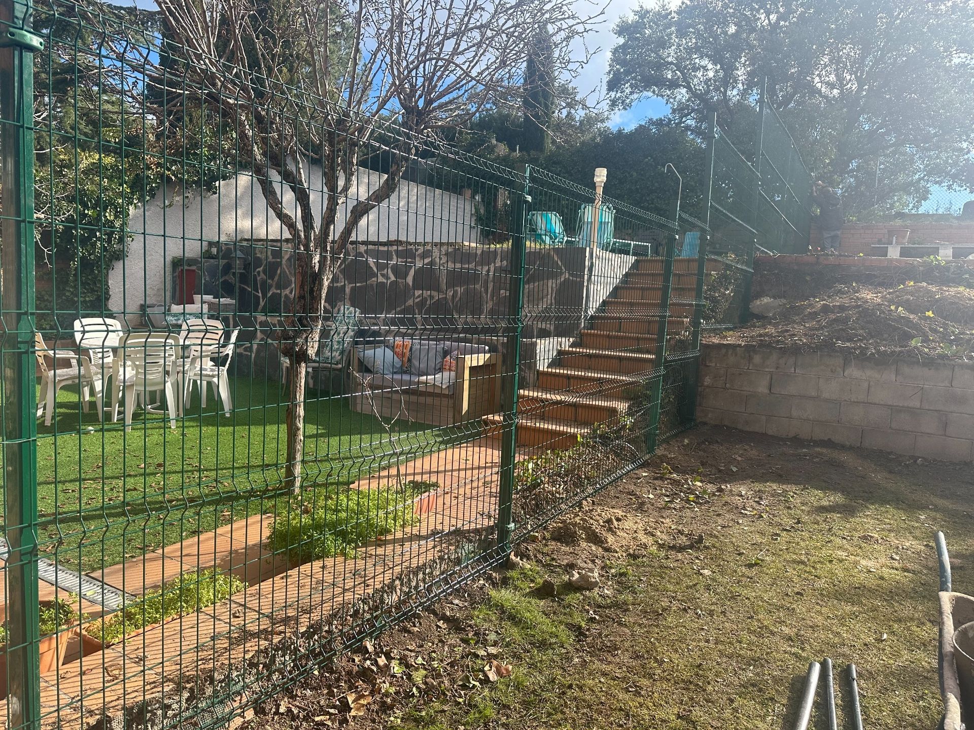 Una cerca de alambre verde rodea un patio trasero con césped, una escalera de ladrillo y un pequeño árbol.