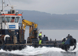 Barco en el agua con grúa. Una persona vestida de amarillo está de pie en la cubierta. Colores azul y blanco.