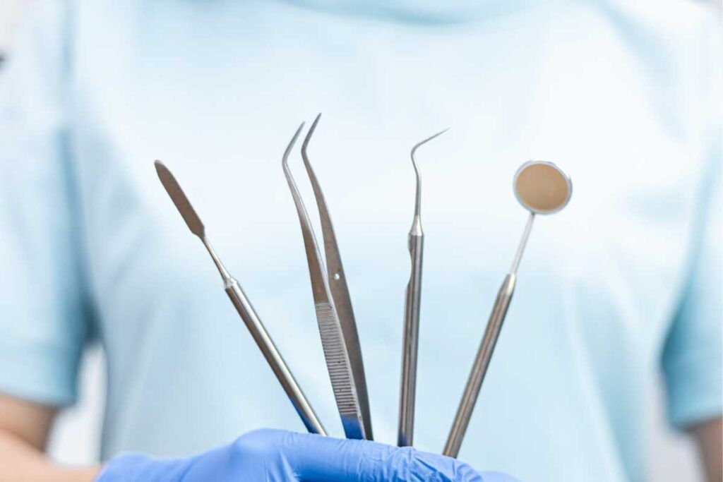 Hammaslääkäri pitelee sinisiin hanskoihin käärityssä kädessään hammaslääketieteellisiä työkaluja, kuten peiliä, anturia ja pihdit.