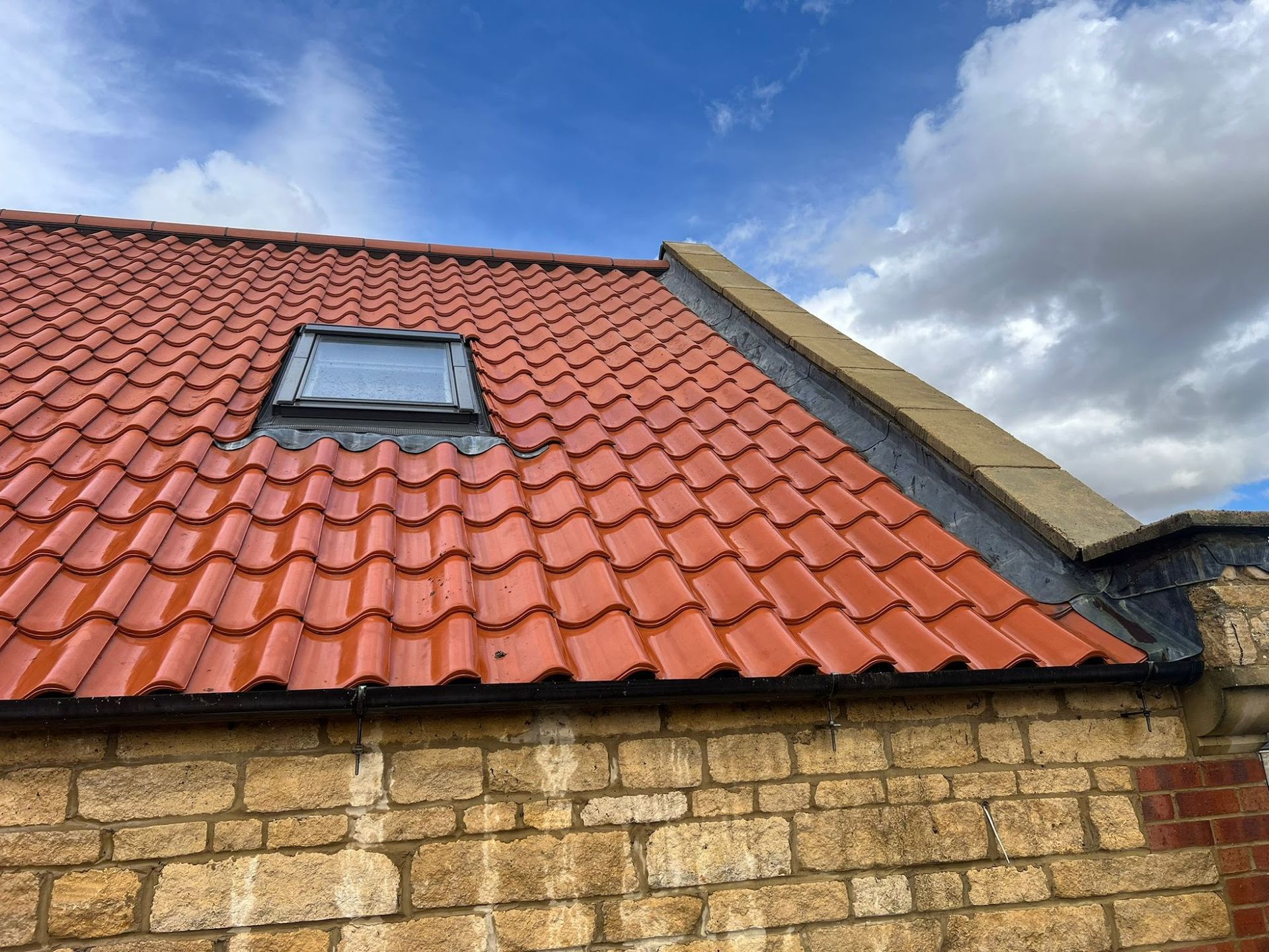 Red tiled roof with a skylight above a stone wall under a cloudy sky