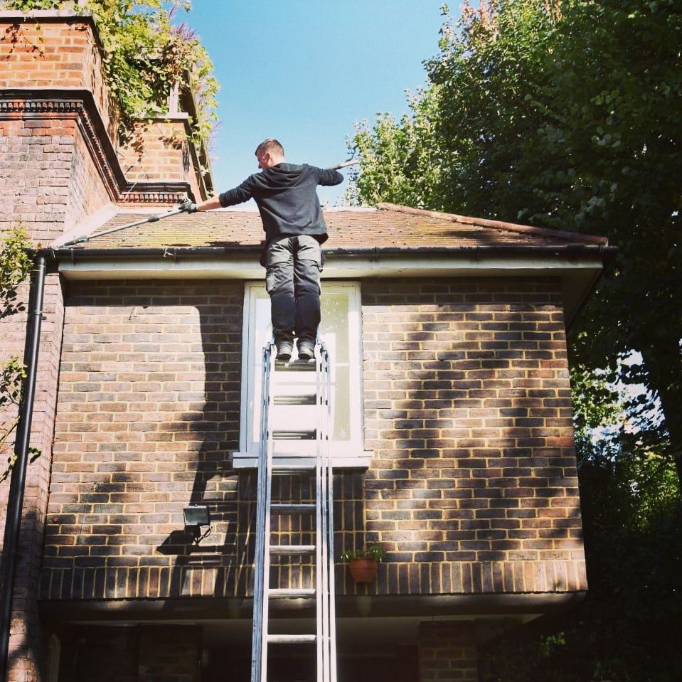 Person standing on a ladder, reaching the roof of a brick house beside a chimney and tree.