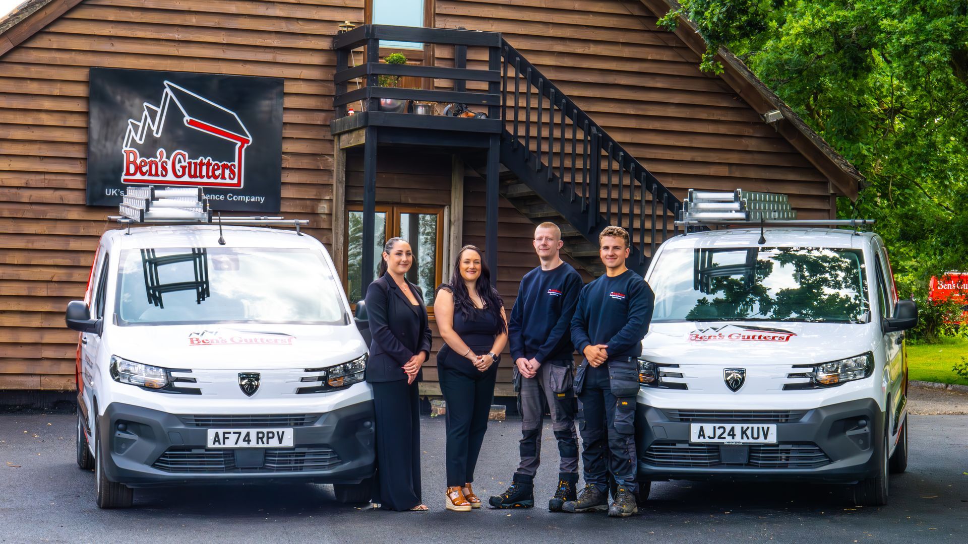 Four people stand between two white vans outside a wooden lodge with a black sign.