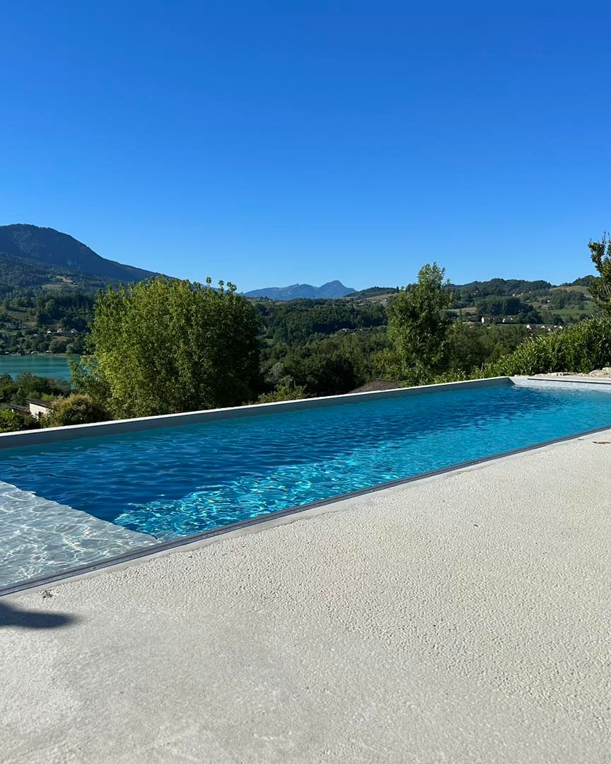 Longue piscine bleue surplombant un lac et des montagnes sous un ciel bleu clair.