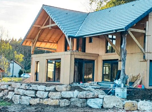 Maison moderne en construction avec poutres apparentes en bois, mur de soutènement en pierre et portes vitrées.