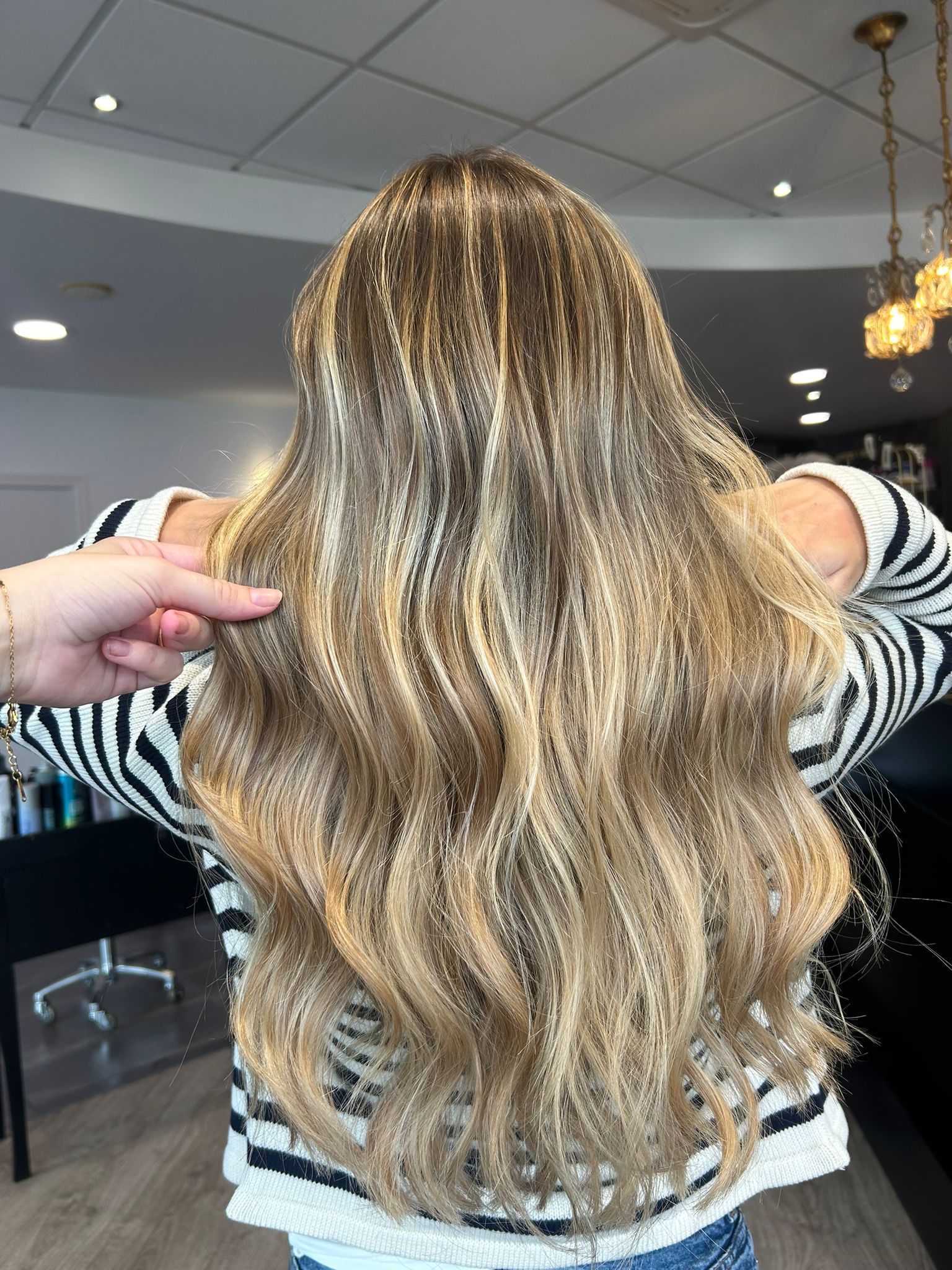 Cheveux bruns ondulés avec des mèches éclaircies. Une main désigne les cheveux. À l'intérieur, elle porte un pull rayé noir et blanc.