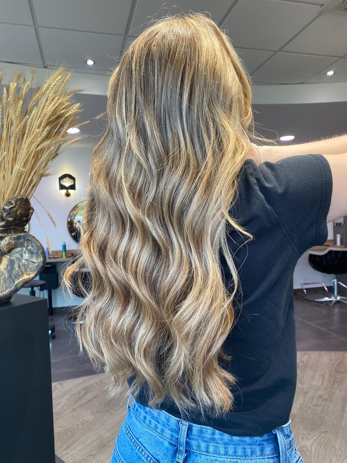 Femme aux longs cheveux ondulés, avec des reflets blonds, portant un t-shirt noir et un jean dans un salon de coiffure.