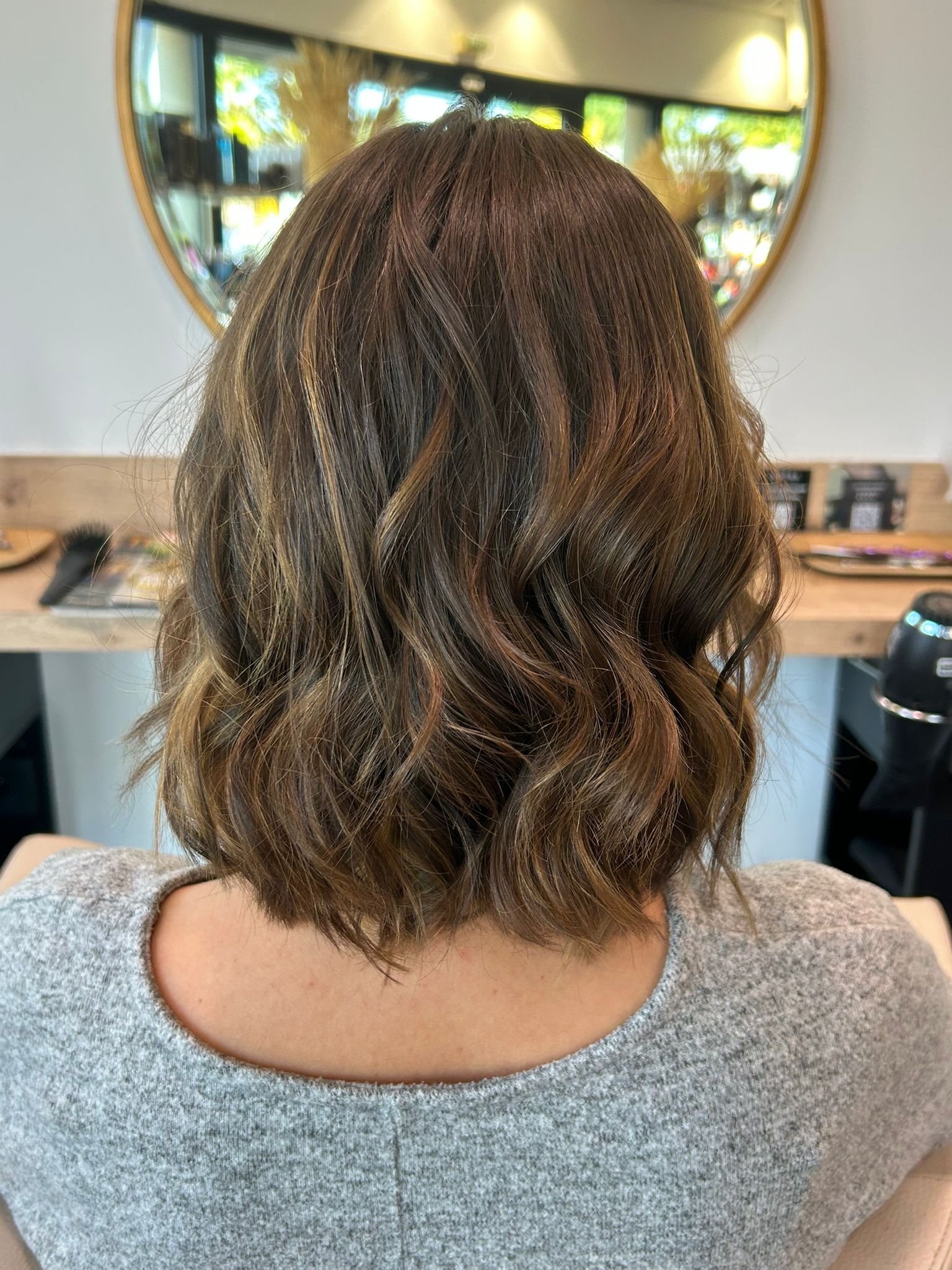 Une femme a une coupe au carré brune et crêpée, vue de dos dans un salon de coiffure, près d'un miroir.