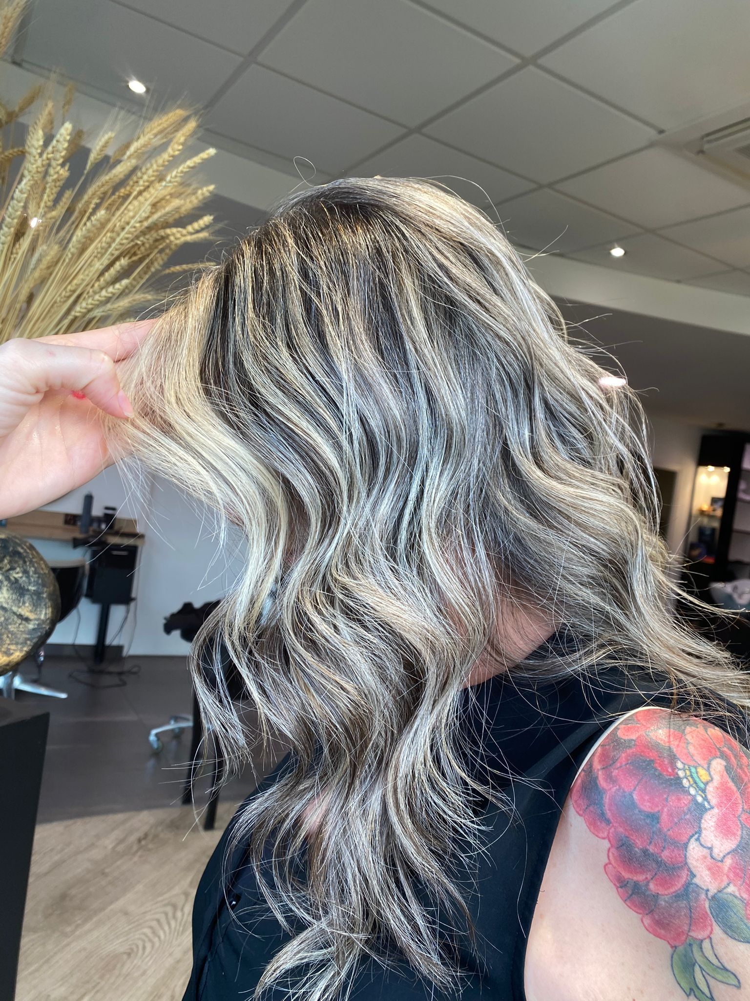 Femme aux cheveux ondulés et méchés ; main touchant ses cheveux dans un salon de coiffure ; un tatouage floral est visible.