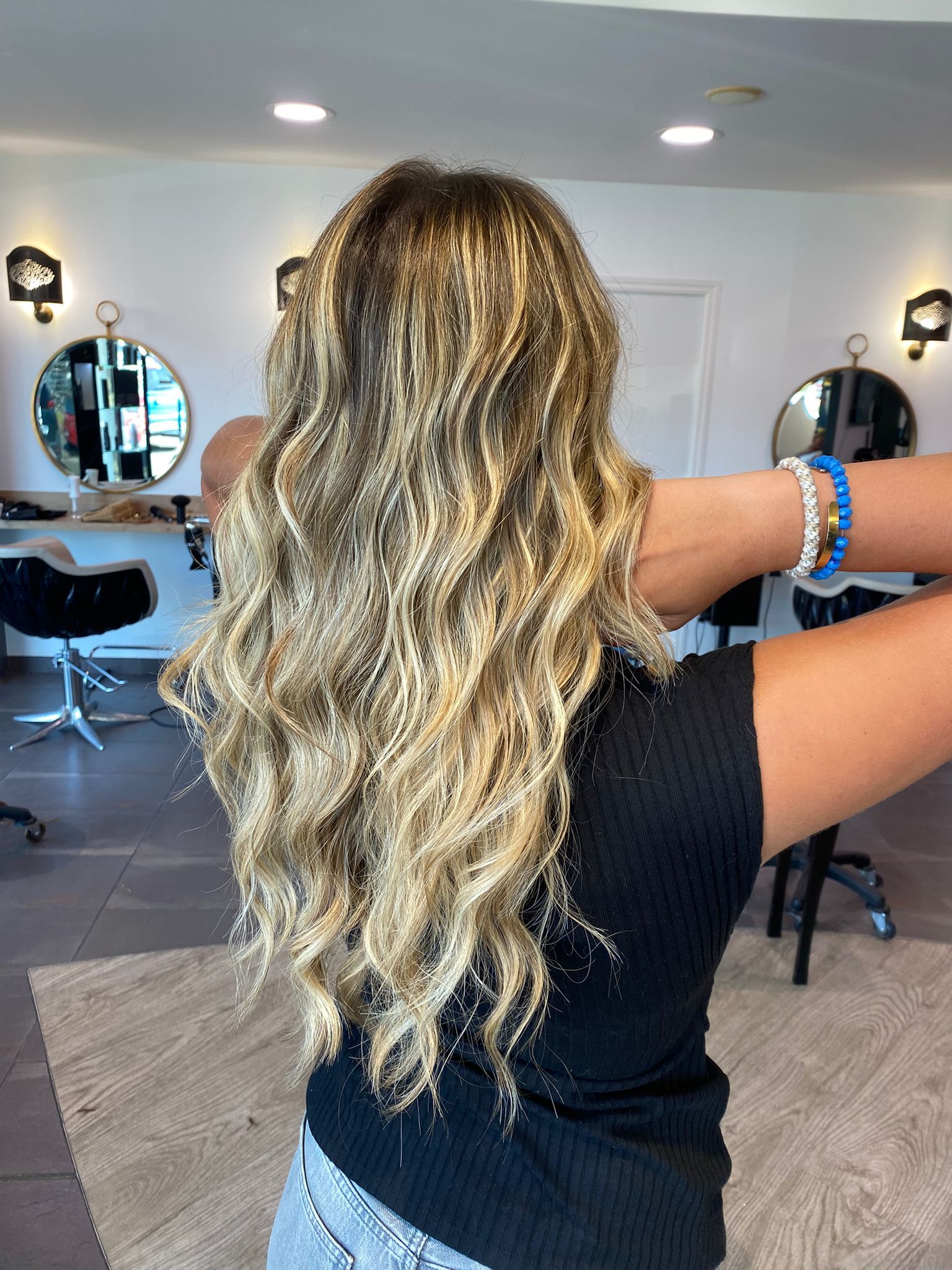 Femme aux longs cheveux blonds ondulés dans un salon de coiffure, les bras écartés.