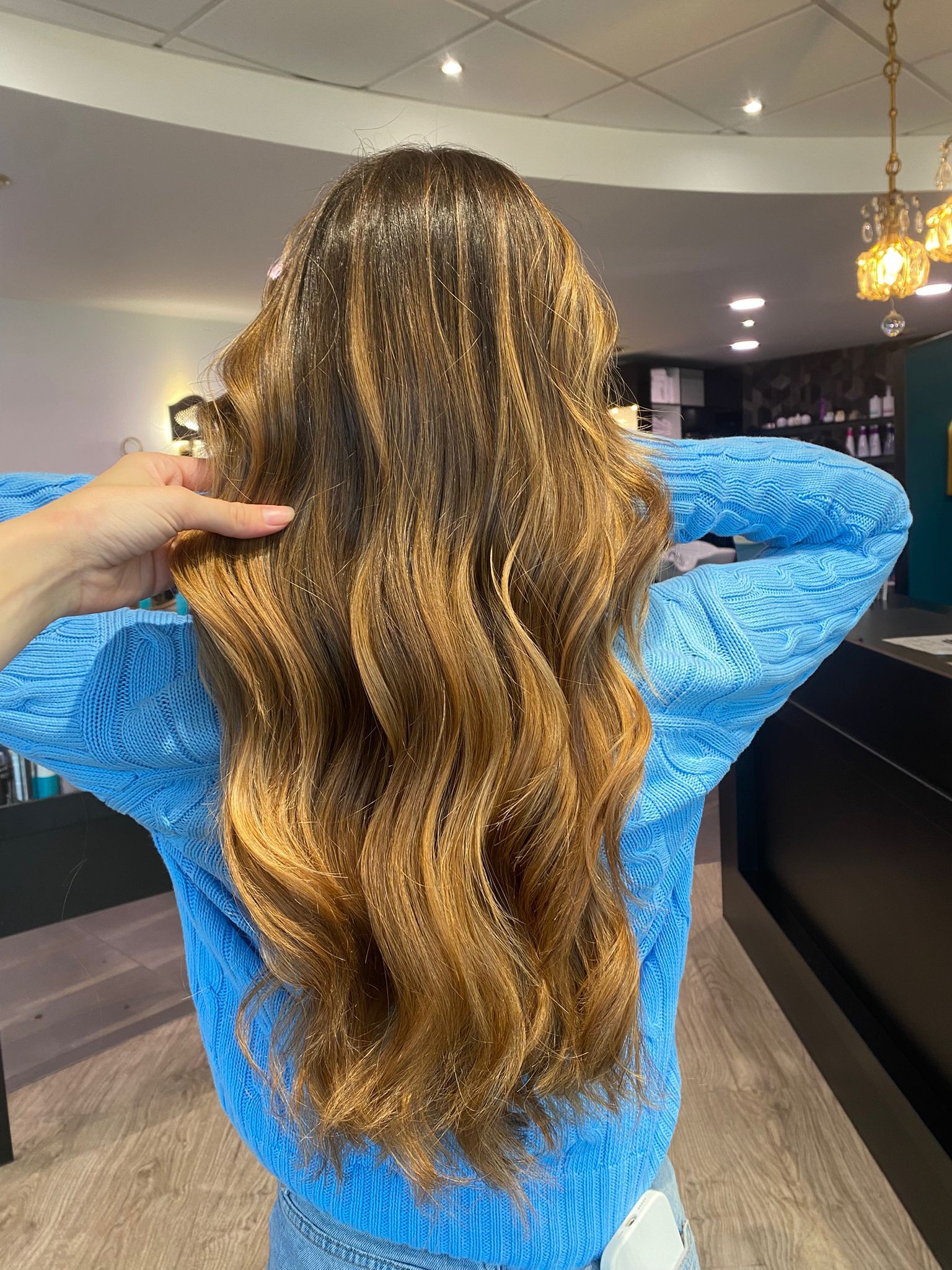 Femme aux cheveux ondulés, avec des mèches brunes, portant un pull bleu, dans un salon de coiffure.