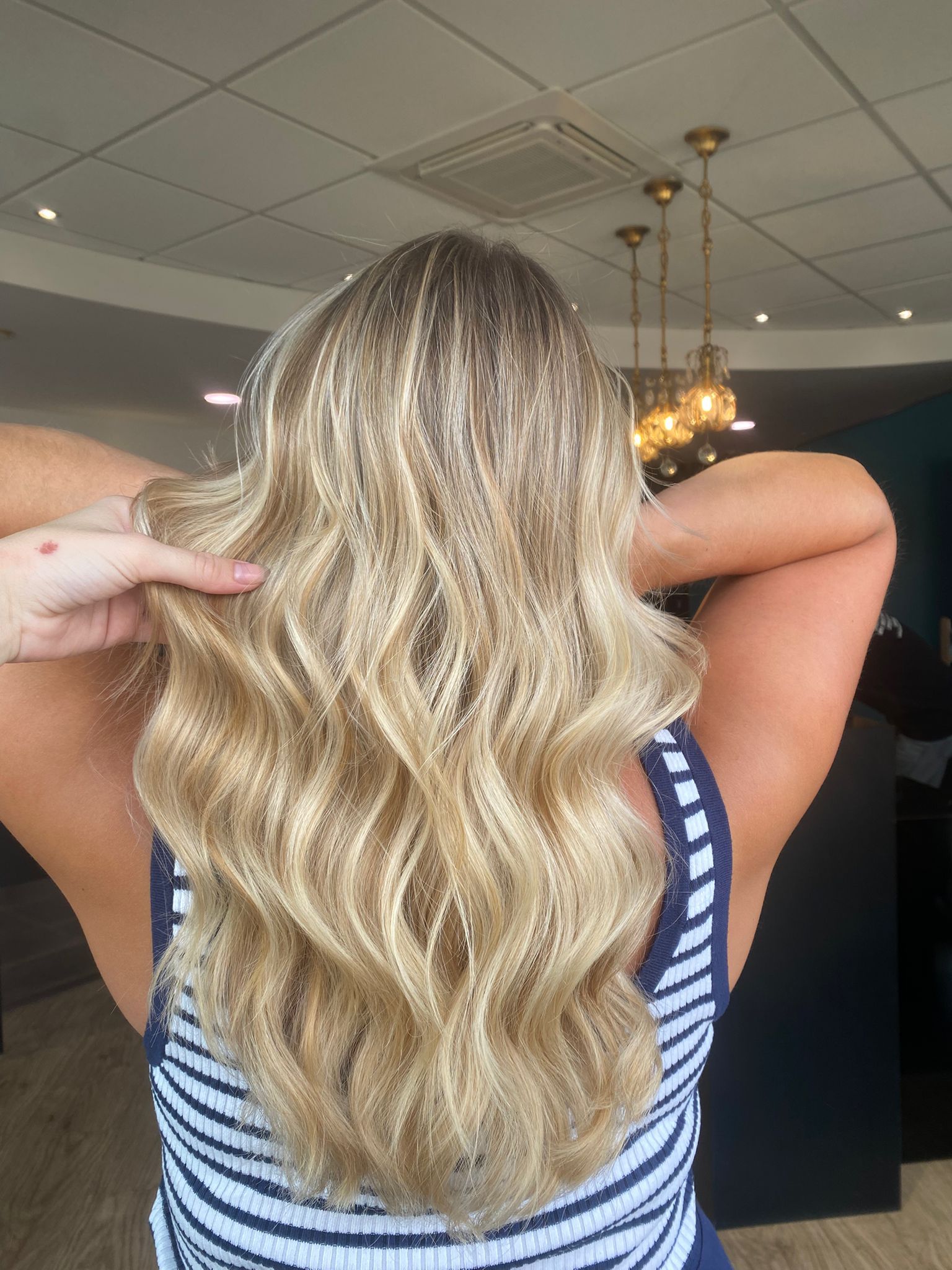Cheveux blonds ondulés, dos d'une personne, main touchant les cheveux. Chemise rayée, ambiance salon avec éclairage.