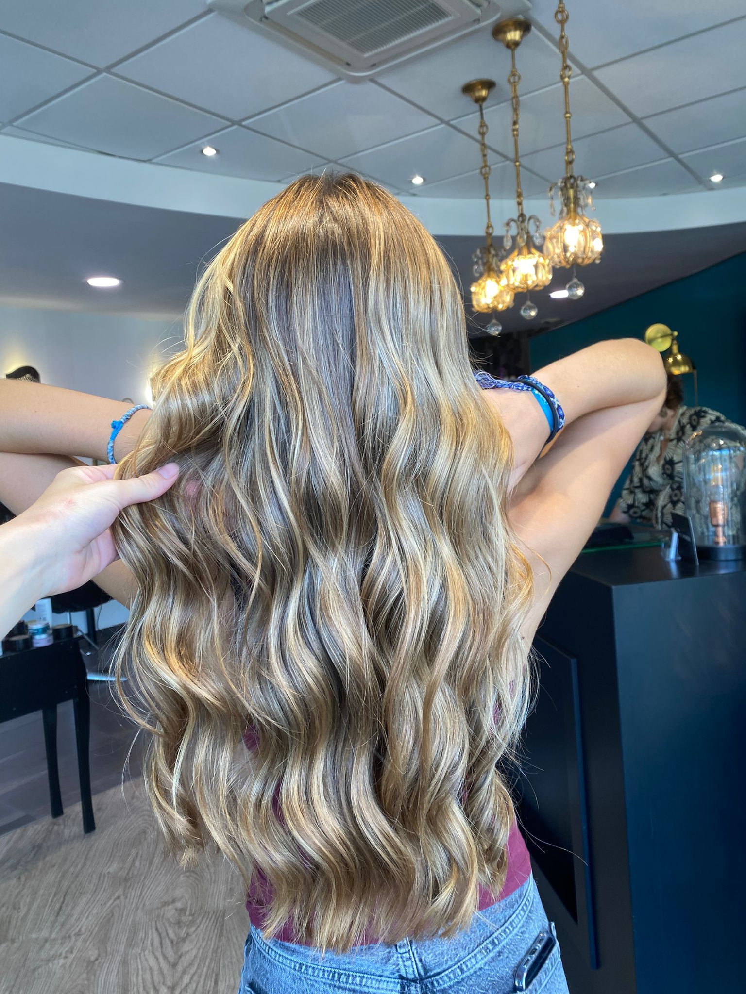 Femme aux longs cheveux blonds ondulés avec des mèches, dans un salon de coiffure, le bras levé, la main touchant ses cheveux.