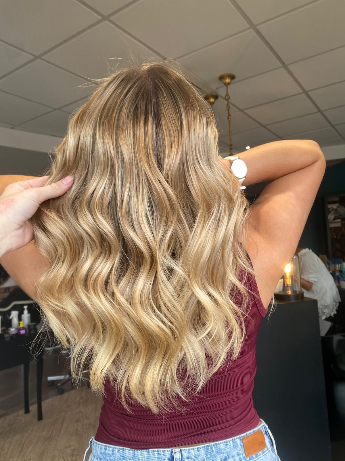 Femme aux cheveux blonds ondulés, portant un haut bordeaux et un jean, dans un salon de coiffure.