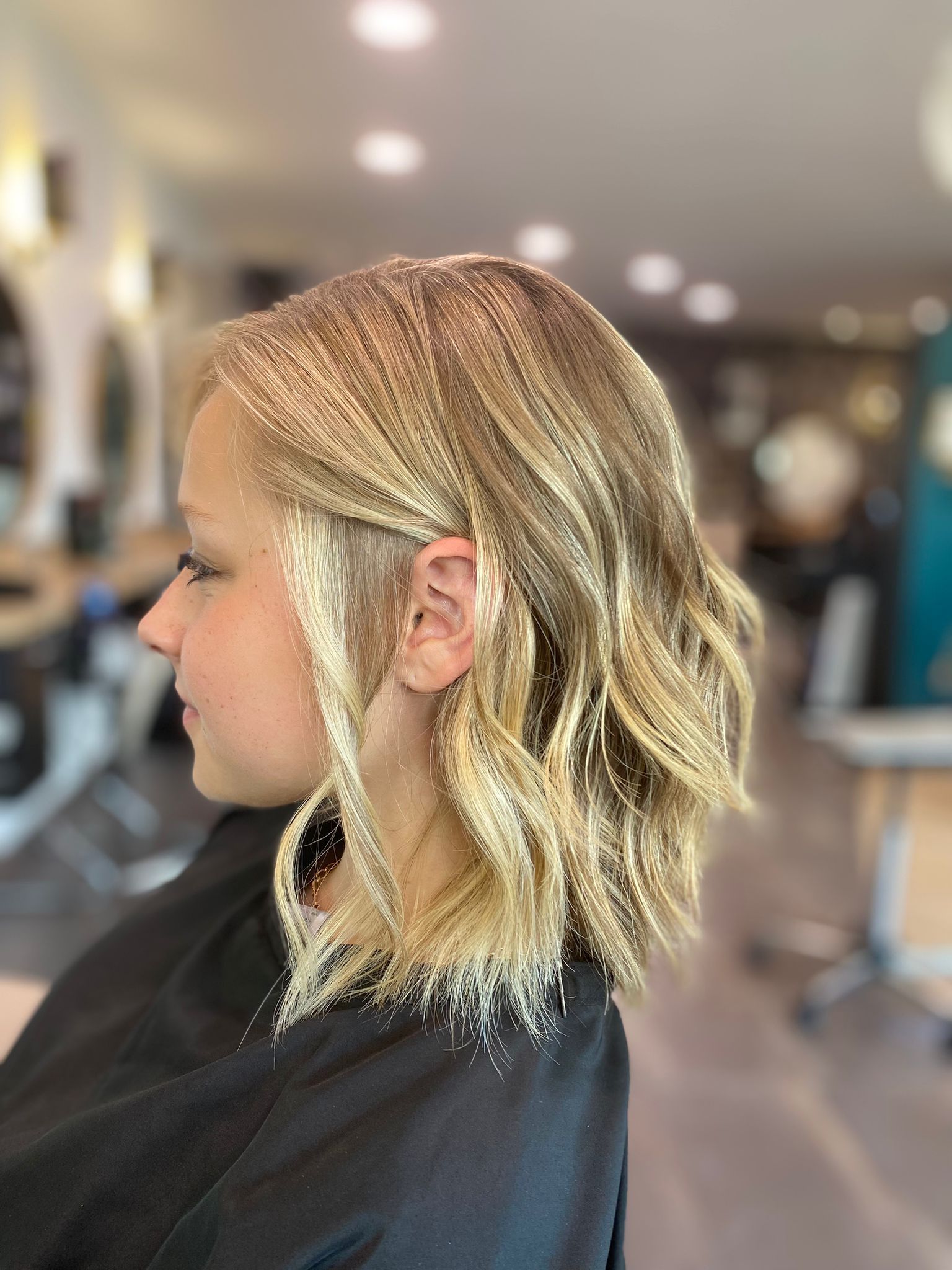 Femme aux cheveux blonds ondulés, coiffée en salon, portant une cape noire.