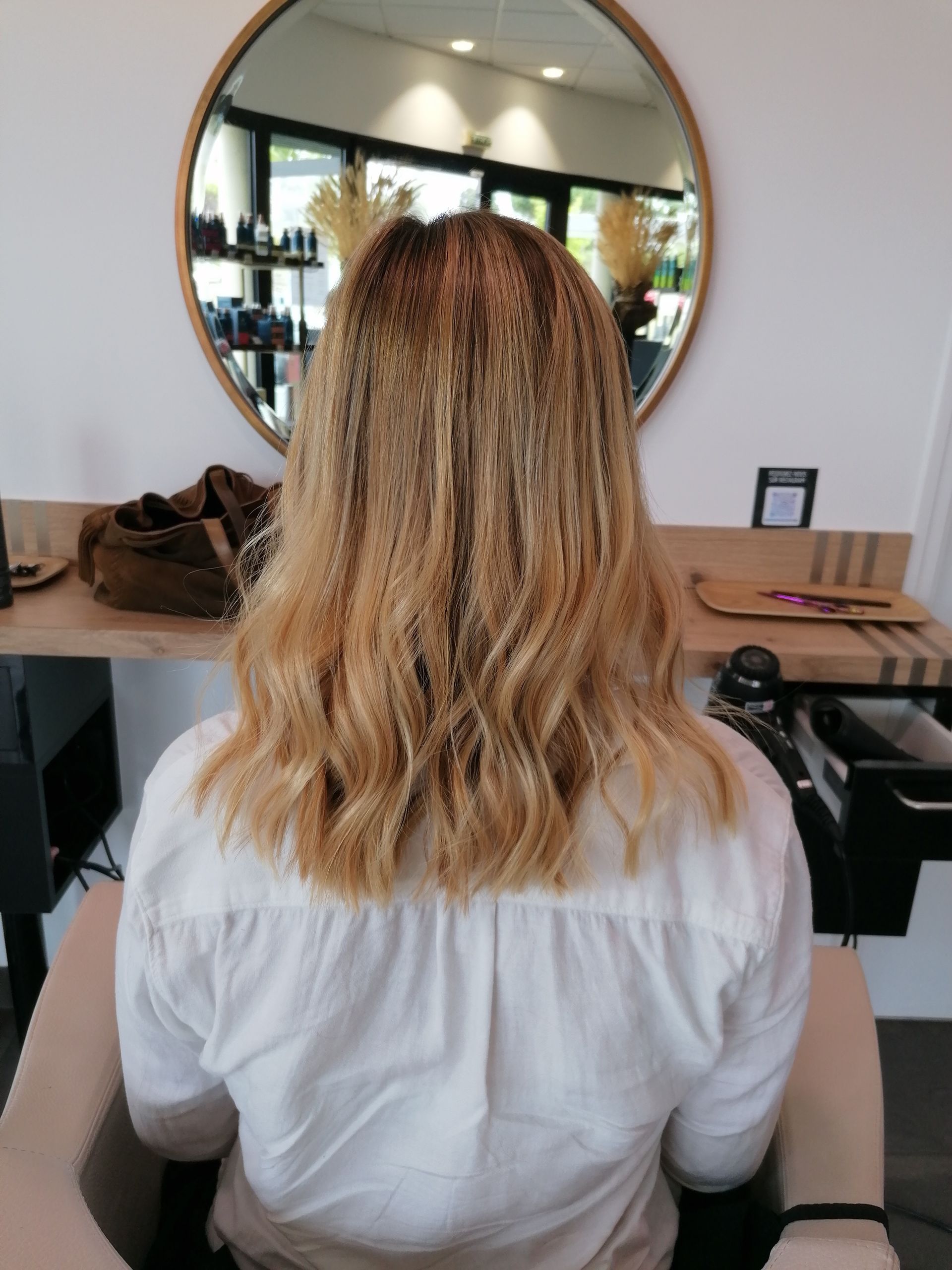 Femme aux cheveux blonds ondulés assise dans un fauteuil de salon, dos à la caméra. Miroir et comptoir en arrière-plan.