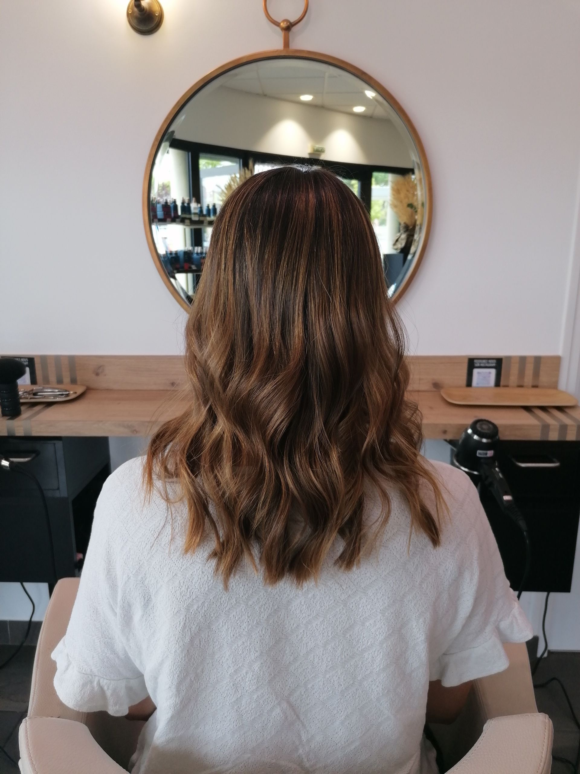 Femme aux cheveux bruns ondulés, assise dans un fauteuil de salon, face à un miroir rond.
