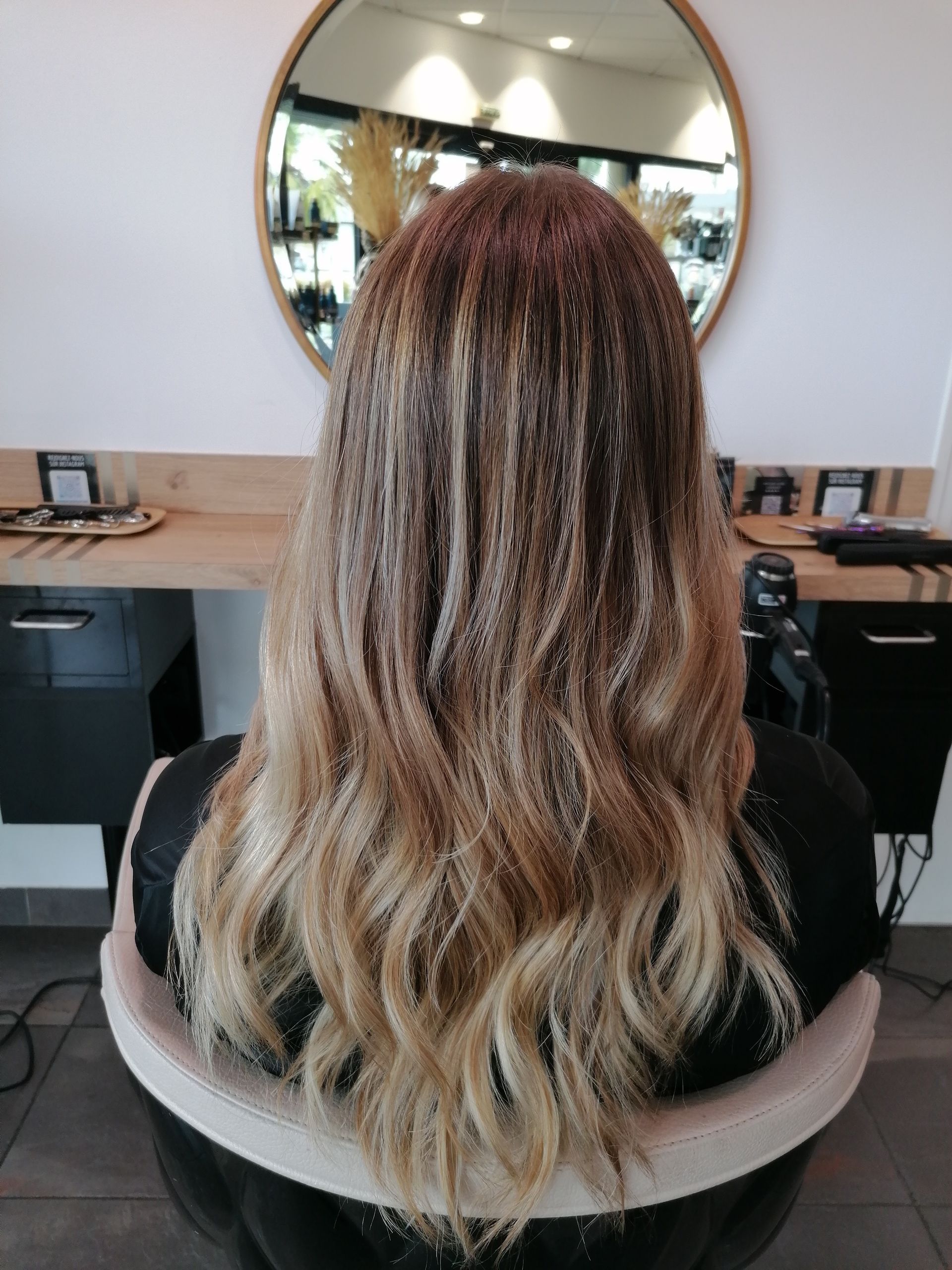 Femme aux longs cheveux ondulés, racines foncées dégradées vers des pointes blondes, assise dans un fauteuil de salon.