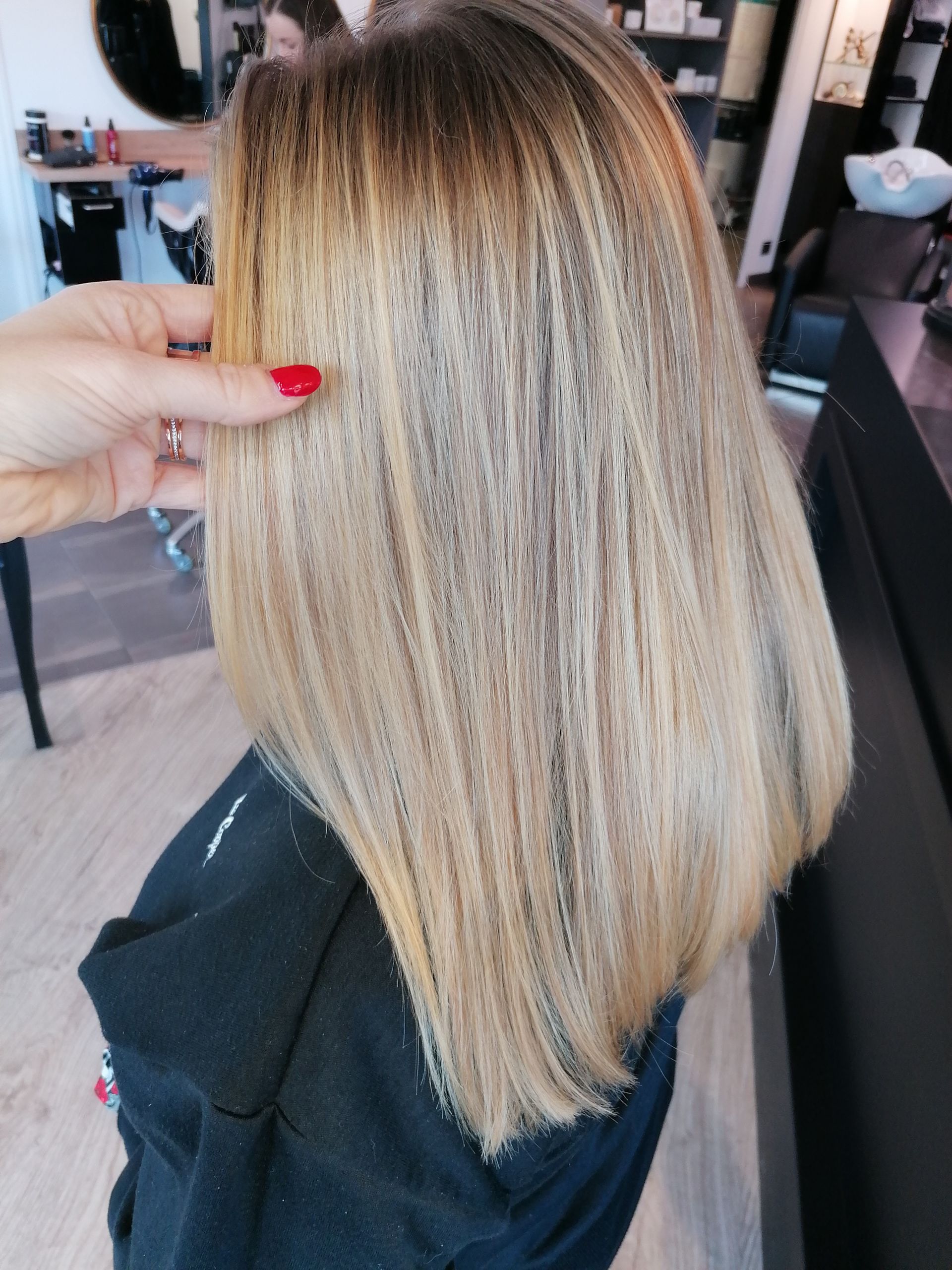 Cheveux blonds aux racines foncées, coiffés en un carré lisse, maintenus par la main d'une personne aux ongles vernis en rouge.