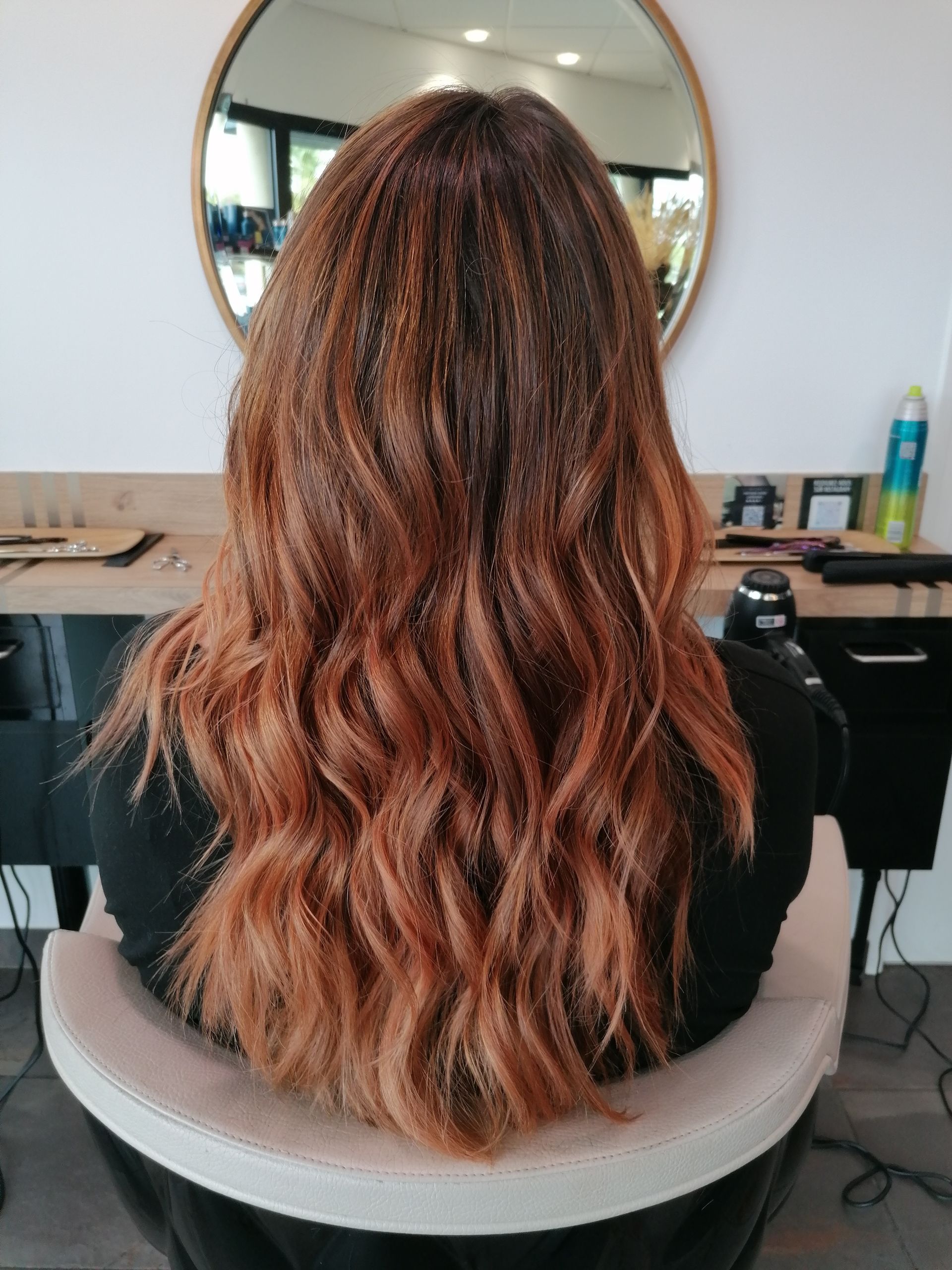 Femme aux longs cheveux ondulés auburn, assise sur un fauteuil de salon, dos à la caméra.