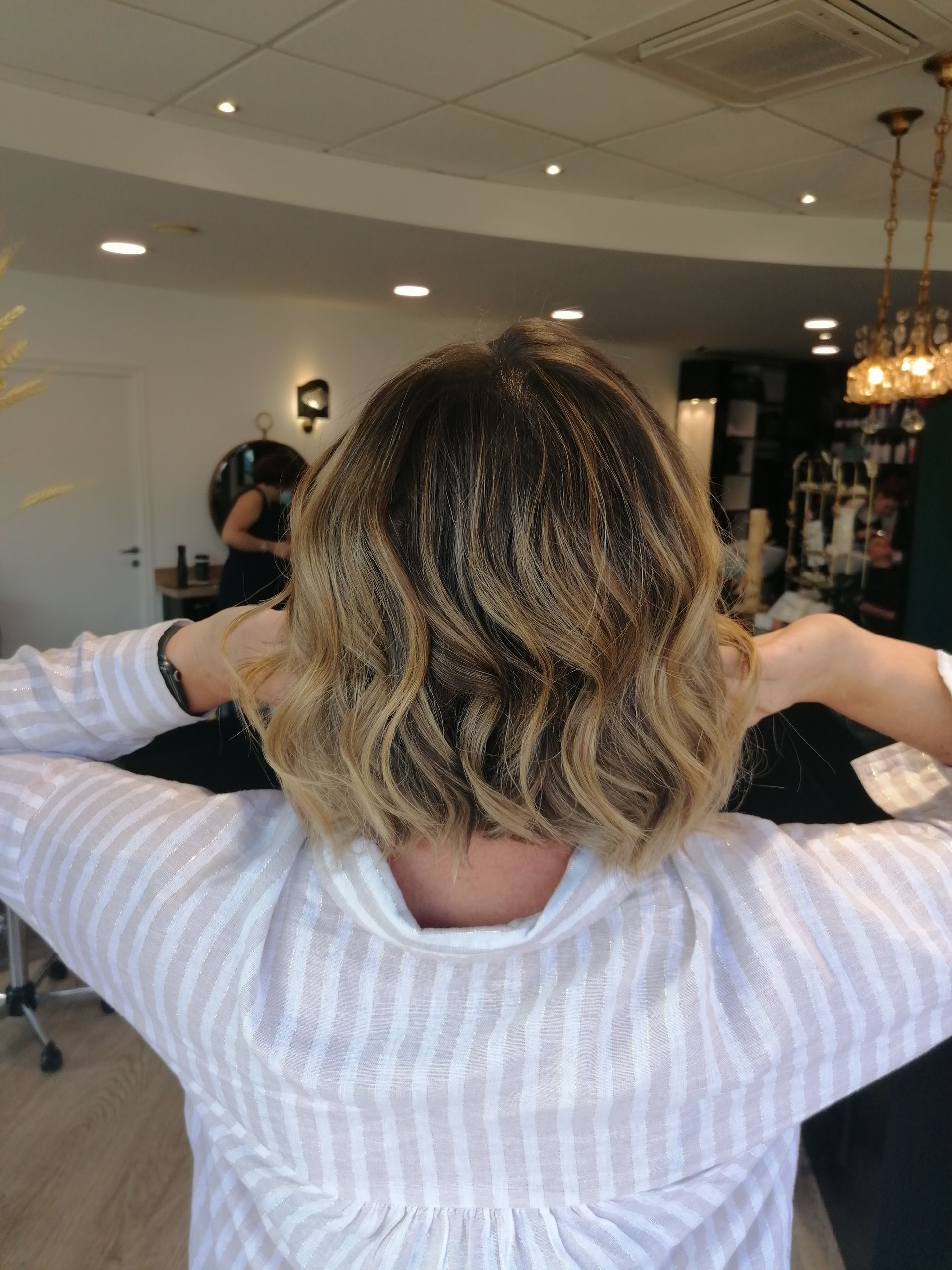 Femme aux cheveux ondulés blonds et bruns, tenant ses cheveux relevés, dans un salon de coiffure.
