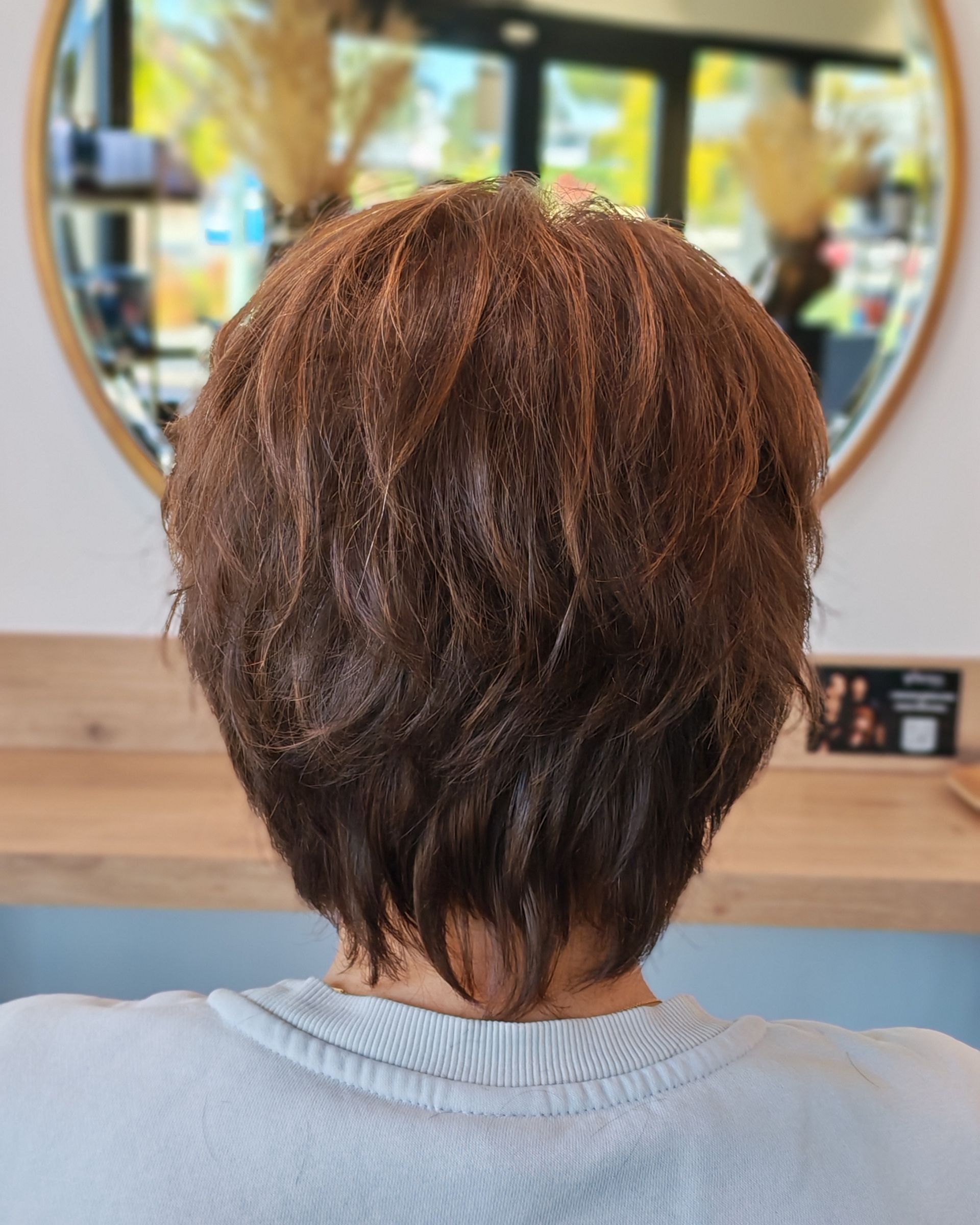 Vue de dos d'une personne aux cheveux bruns courts et dégradés, dans un salon de coiffure, se reflétant dans un miroir rond.