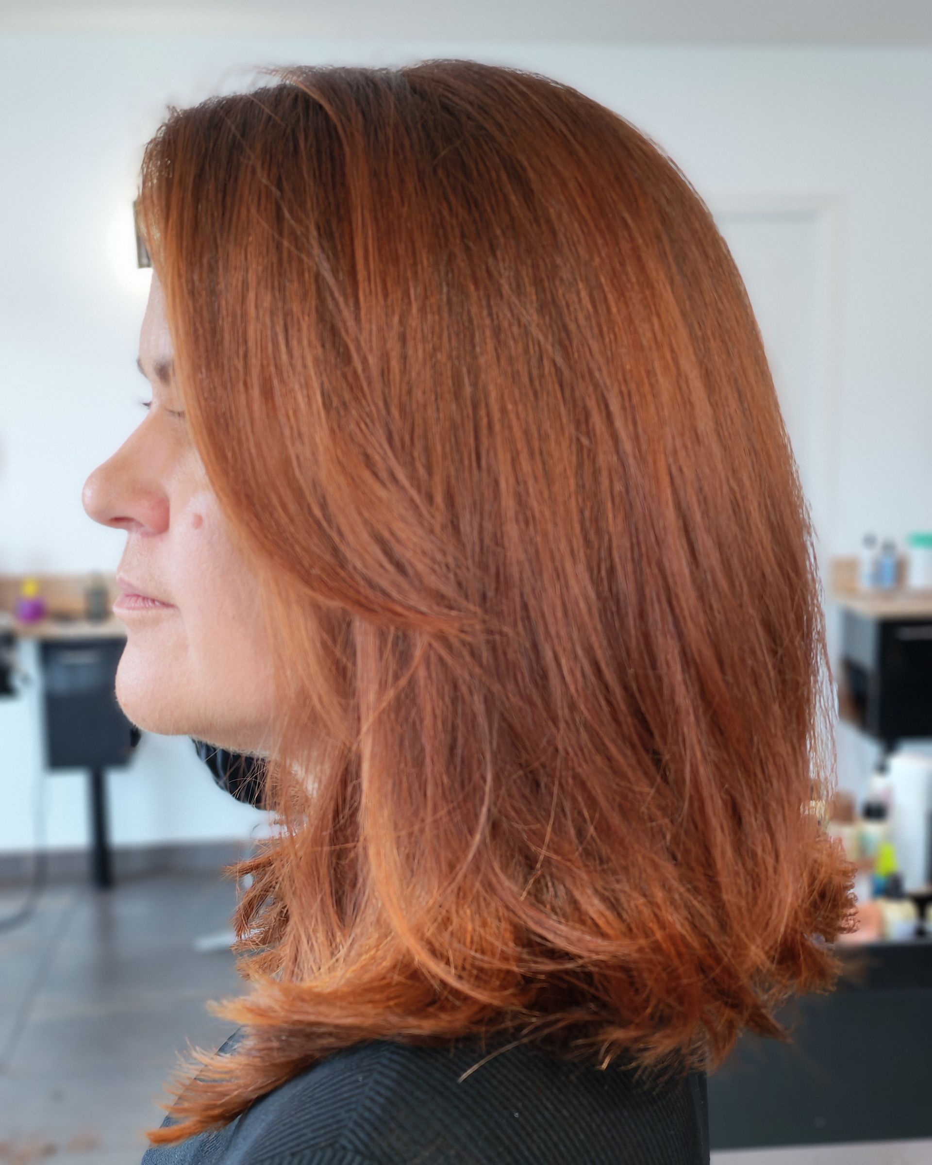 Femme rousse, de profil, dans un salon de coiffure. Cheveux dégradés, mi-longs, d'un roux chaud.