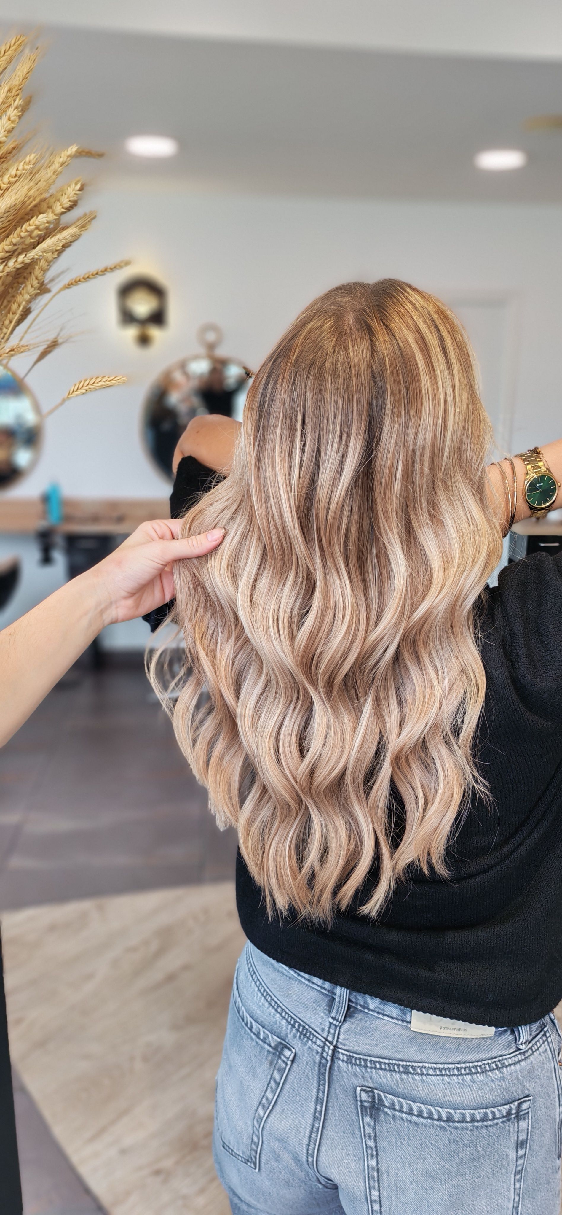 Une personne aux cheveux blonds ondulés se trouve dans un salon de coiffure. Une main caresse ses cheveux, avec des éléments dorés et des miroirs en arrière-plan.