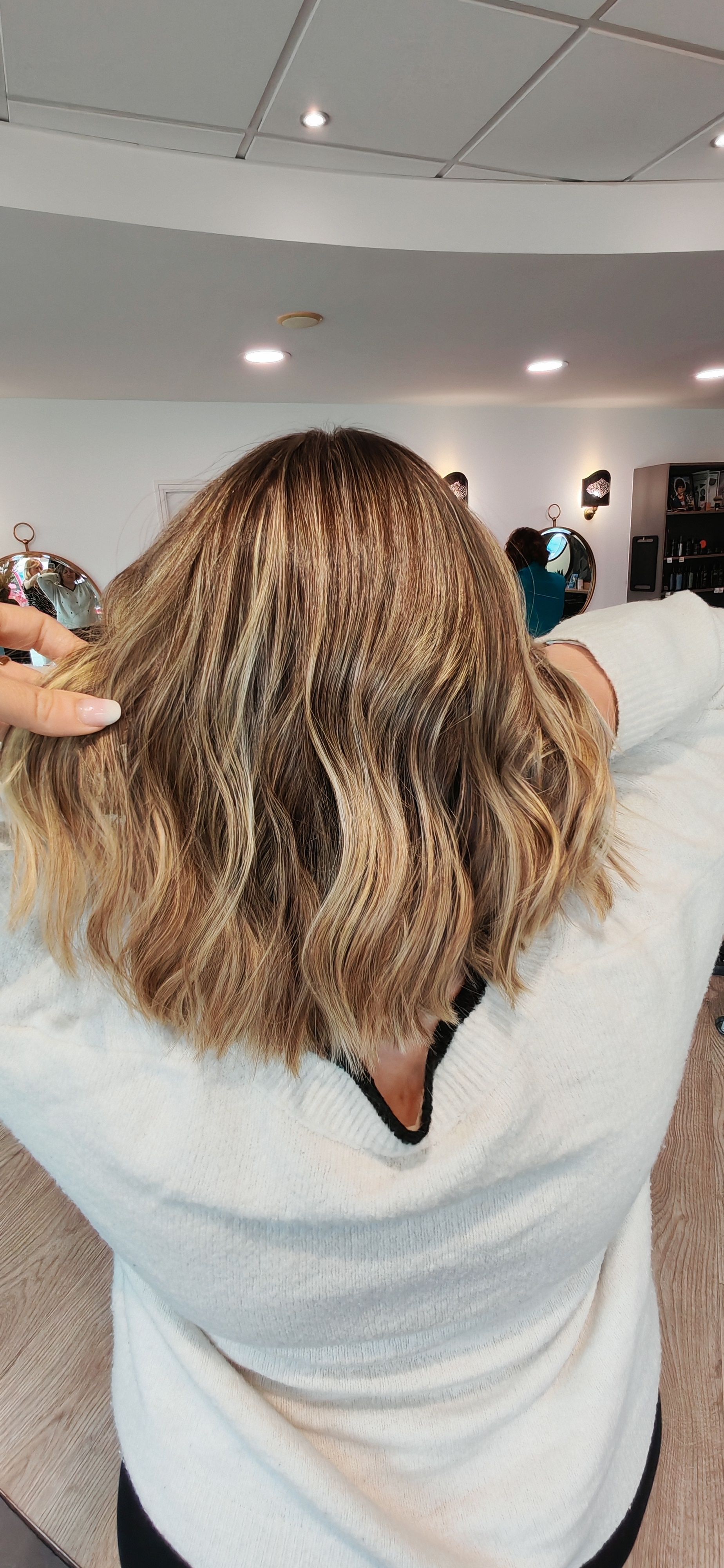 Personne aux cheveux ondulés et méchés, portant un pull blanc, regardant l'objectif. Ambiance salon de coiffure.