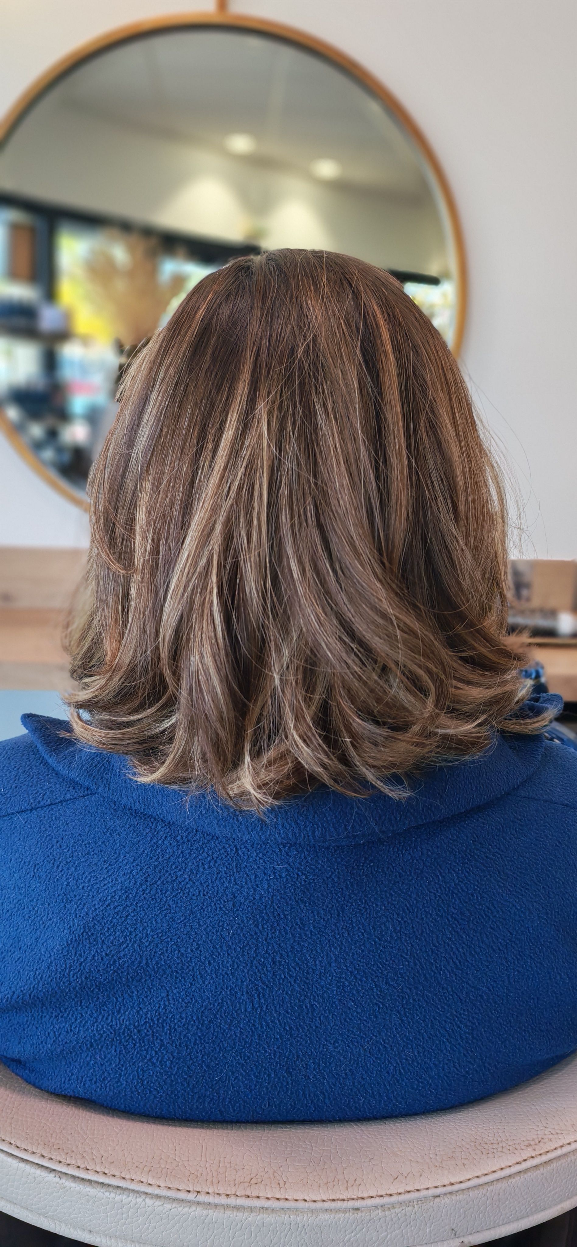 Personne aux cheveux bruns bouclés, dos à la caméra, dans un salon de coiffure, portant un pull bleu.