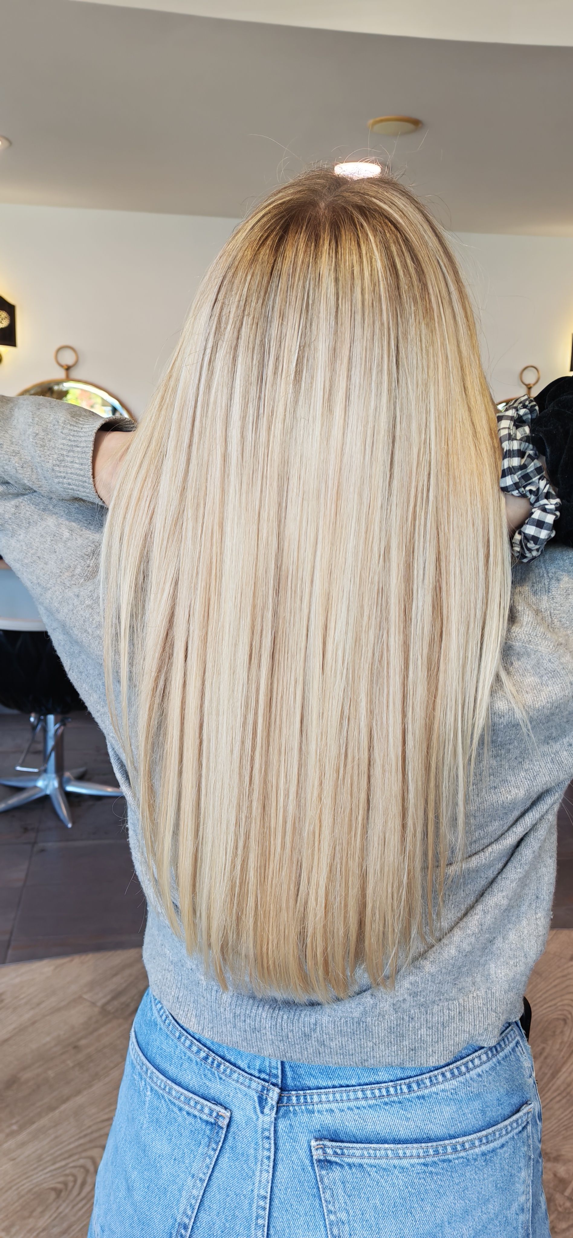 Femme aux longs cheveux blonds ondulés, portant un pull gris et un jean bleu.