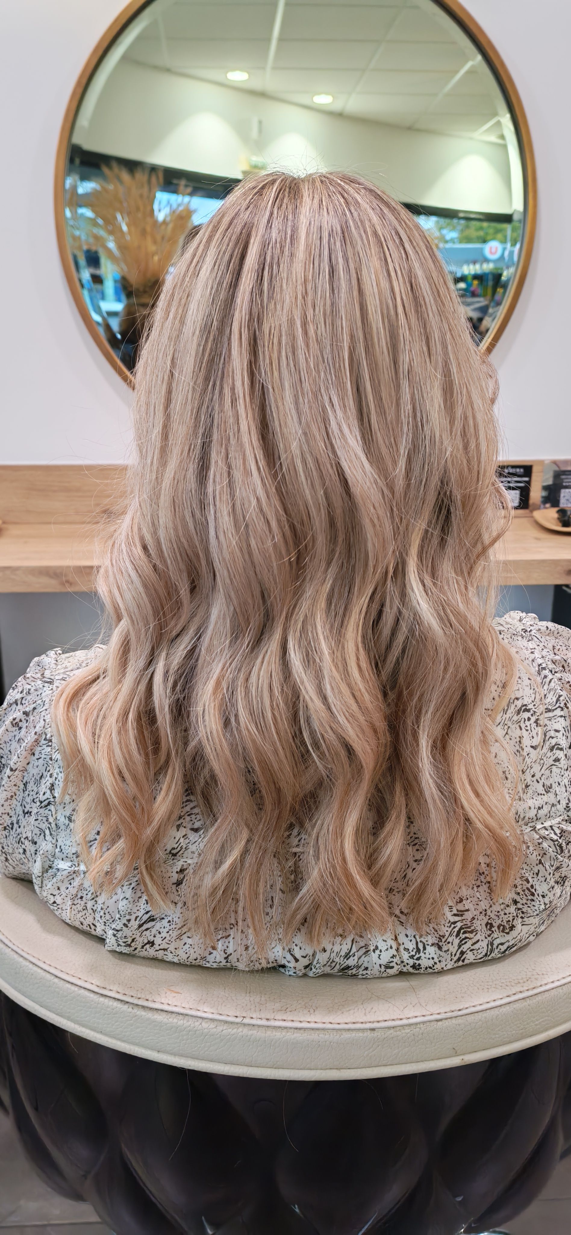 Cheveux blonds ondulés vus de dos, dans un salon de coiffure. Miroir rond doré en arrière-plan.