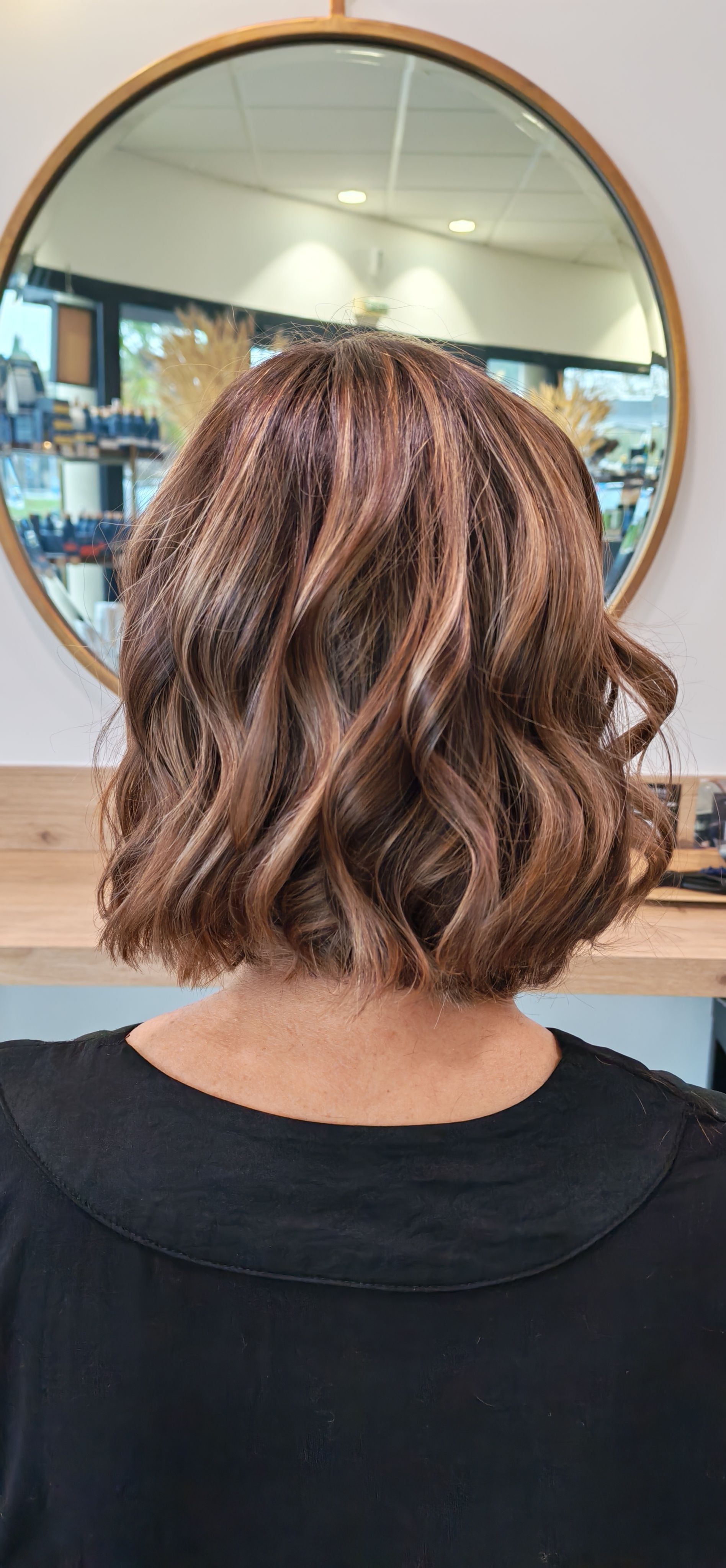Une personne aux cheveux bruns ondulés avec des mèches, se tenant devant un miroir dans un salon de coiffure.