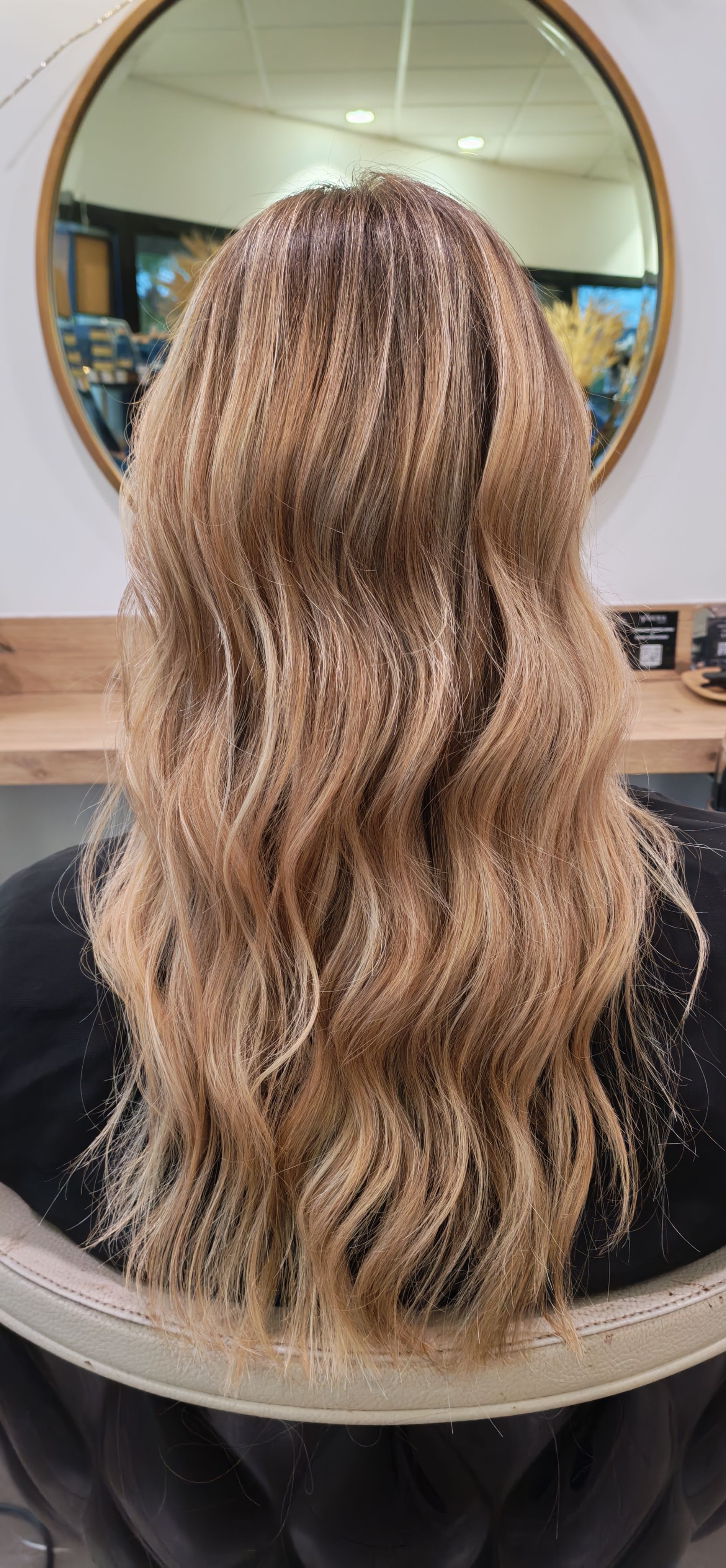 Femme aux longs cheveux blonds ondulés dans un salon de coiffure, vue de dos. Elle est assise, un grand miroir derrière elle.