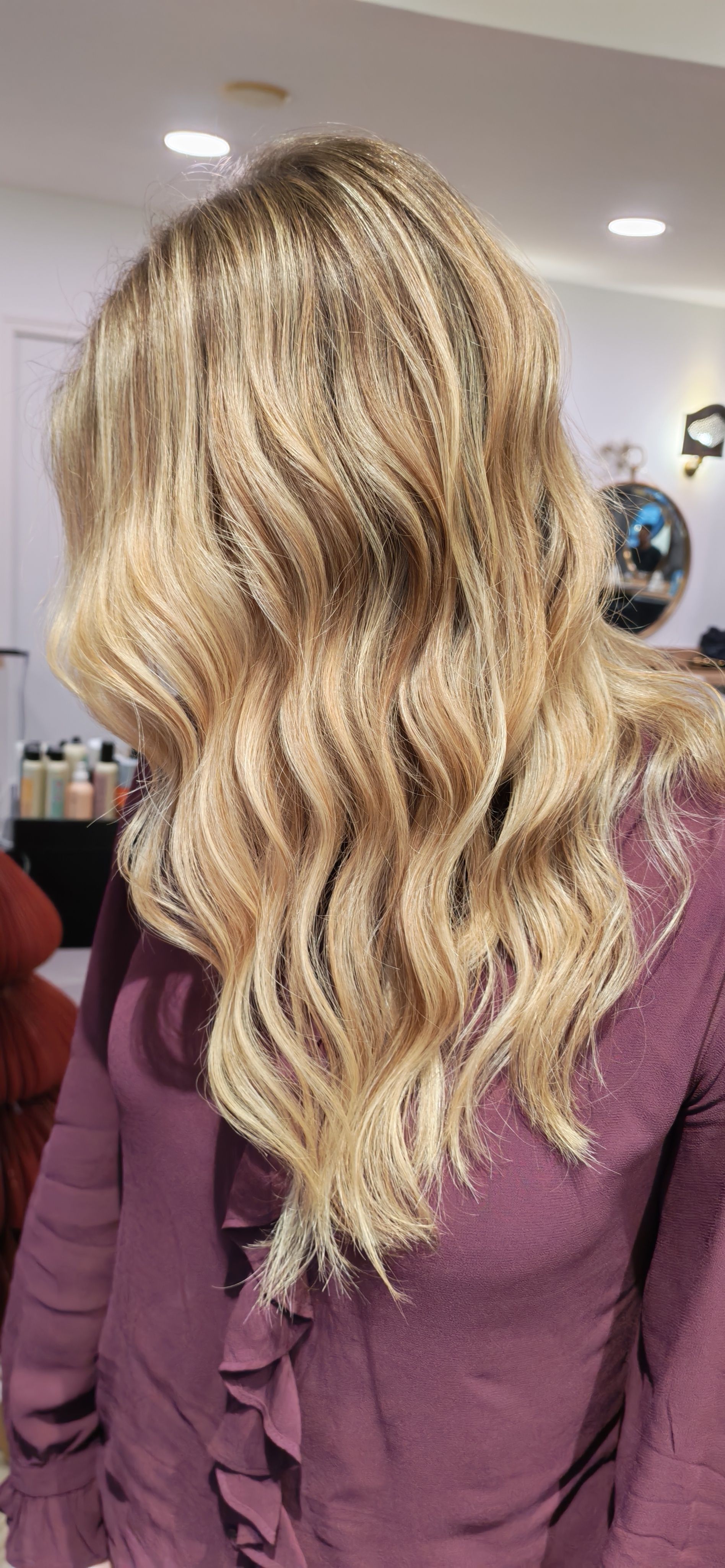 Femme aux cheveux blonds ondulés, portant un chemisier violet à volants, dans un salon bien éclairé.