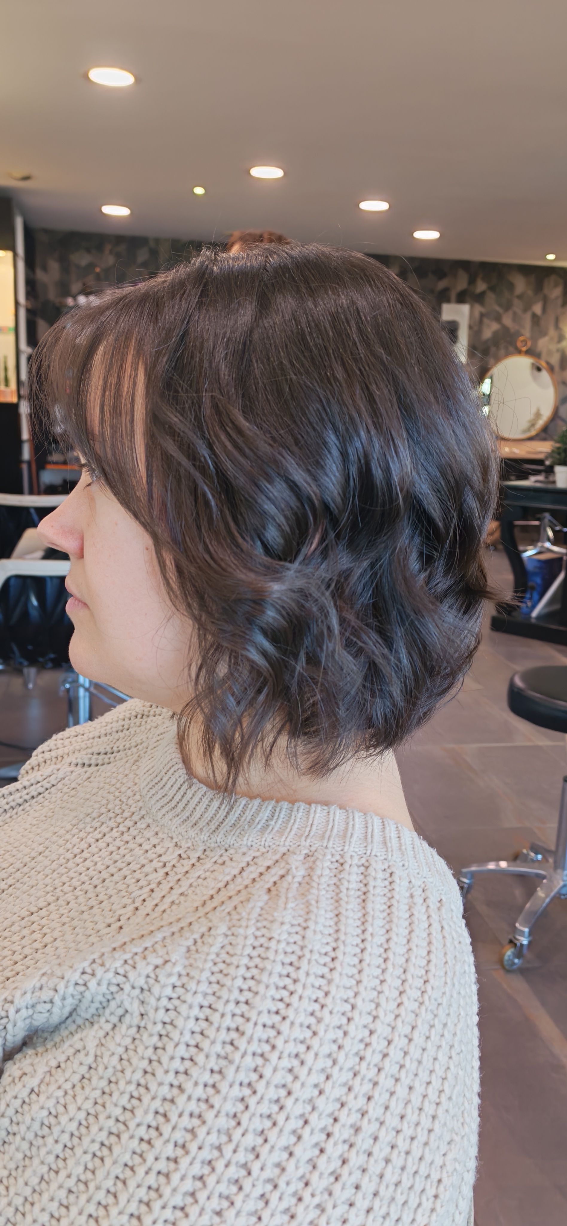 Femme aux cheveux courts, foncés et dégradés, assise dans un salon de coiffure, regardant sur le côté. Elle porte un pull clair.