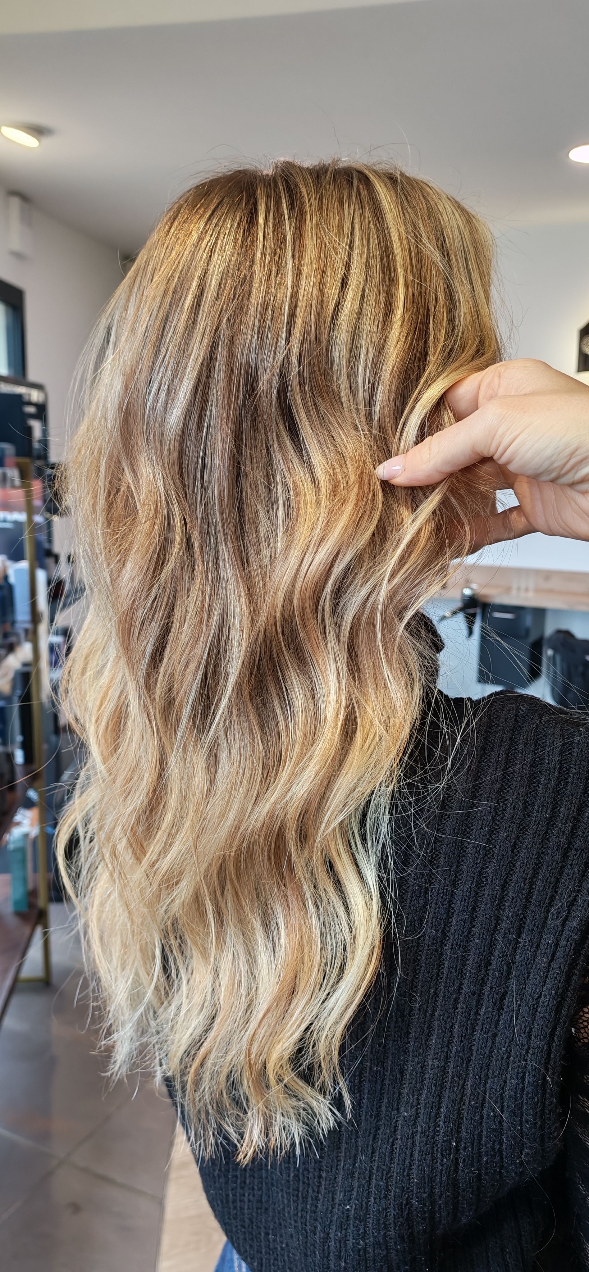 Cheveux blonds et bouclés d'une femme ; une main caresse ses cheveux. Elle porte un pull noir en dessous.