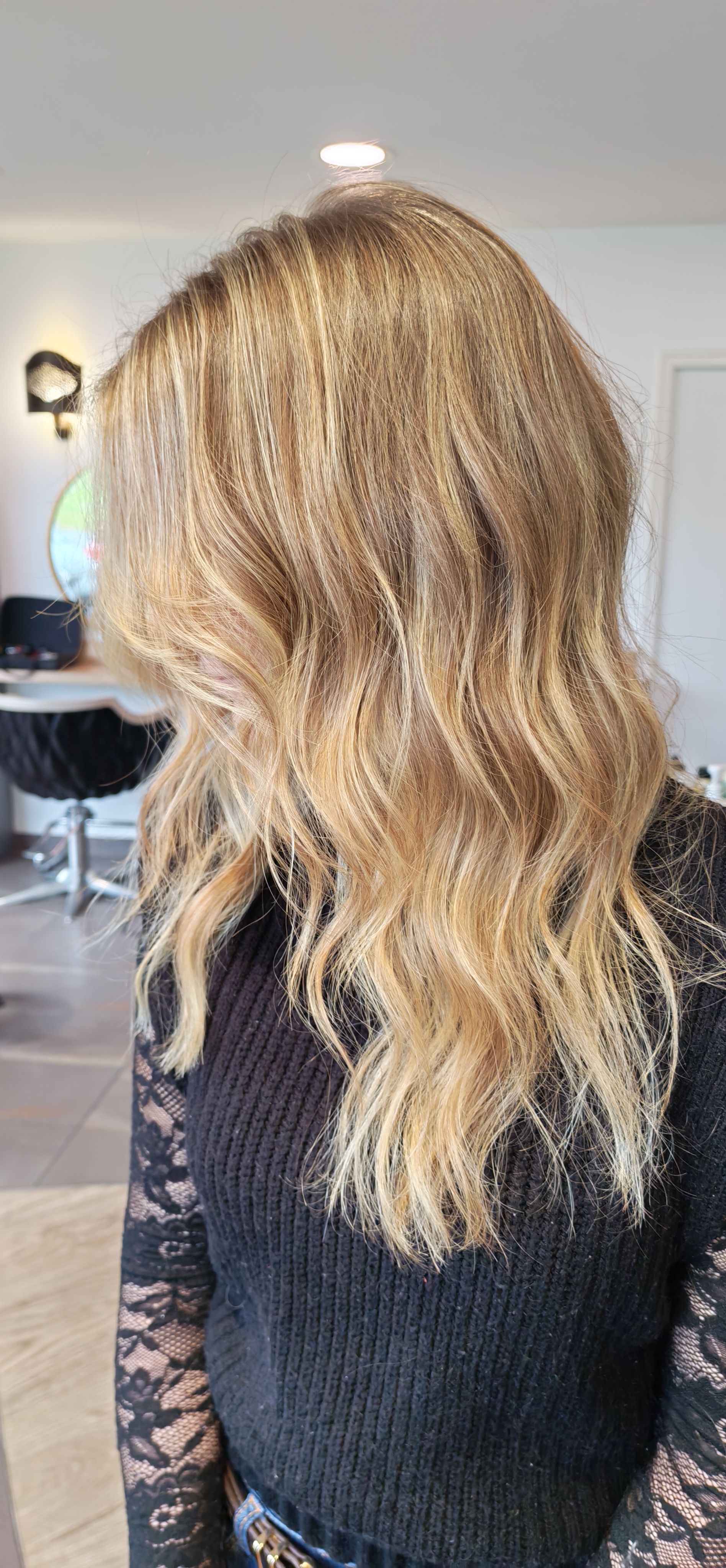 Une personne aux longs cheveux blonds ondulés, portant un pull noir à manches en dentelle, dans un salon de coiffure.