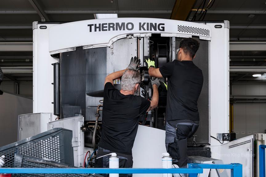 Un homme sur une échelle, au sommet d'un camion orange, inspecte le toit d'une remorque frigorifique blanche ; un camion rouge est visible en arrière-plan.