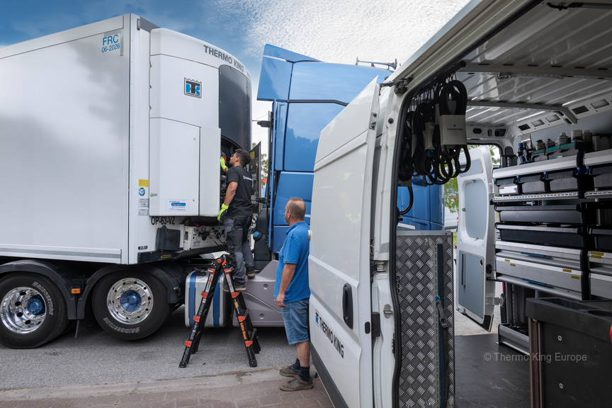 Un homme sur une échelle, au sommet d'un camion orange, inspecte le toit d'une remorque frigorifique blanche ; un camion rouge est visible en arrière-plan.