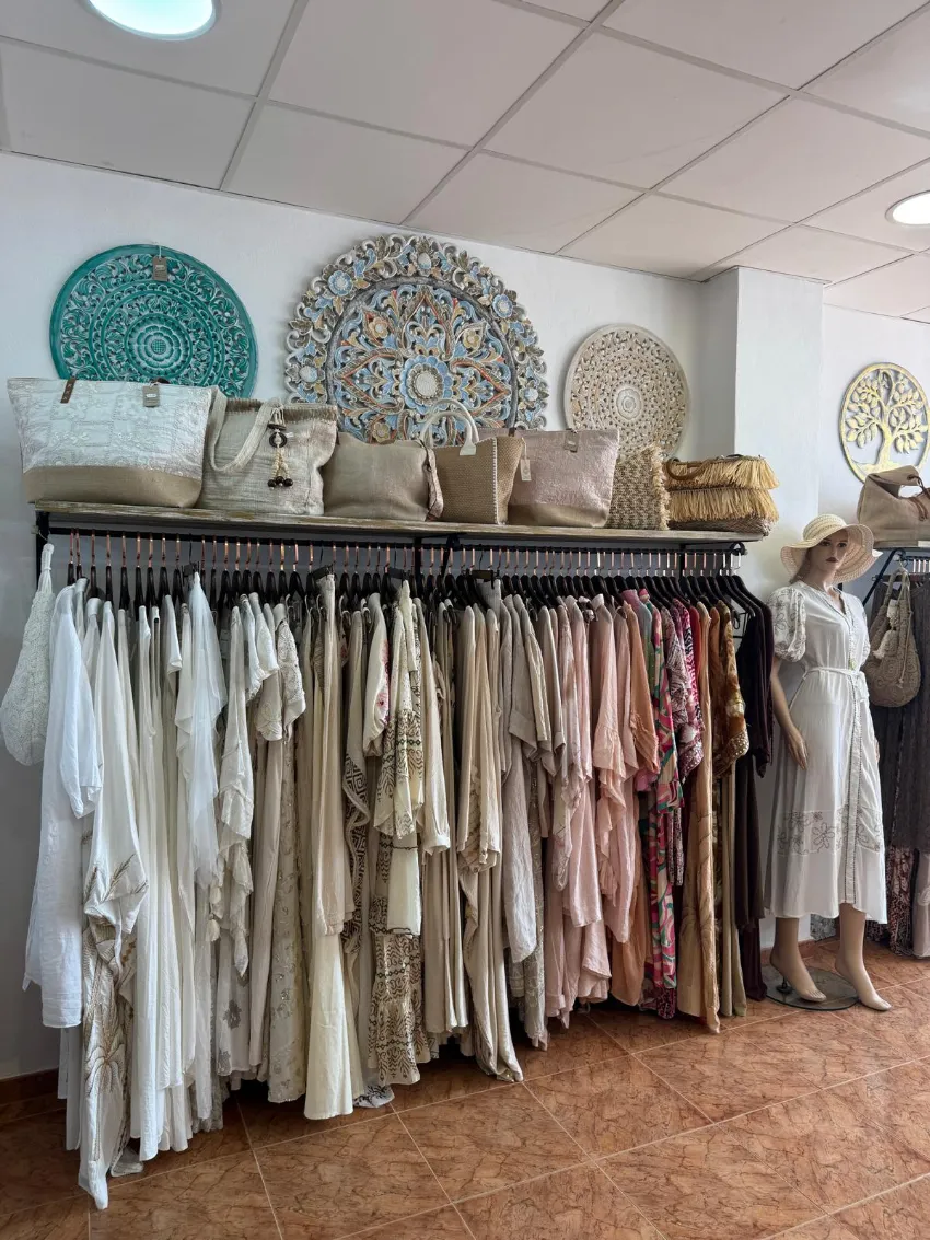 Un perchero de boutique con vestidos en tonos neutros y bolsos tejidos, arte decorativo en la pared y un maniquí a la derecha.
