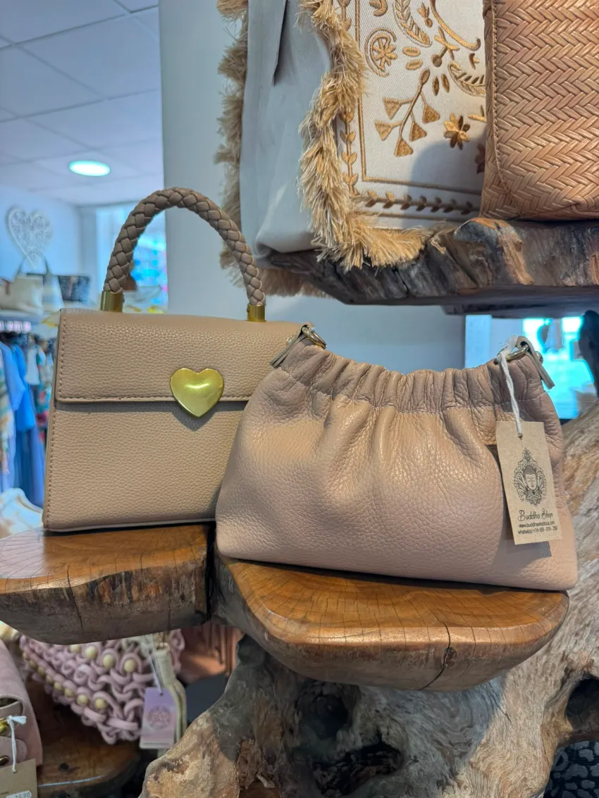 Dos bolsos sobre pedestales de madera rústica en una boutique, uno de color tostado con un cierre de corazón verde y otro beige con cordón ajustable.