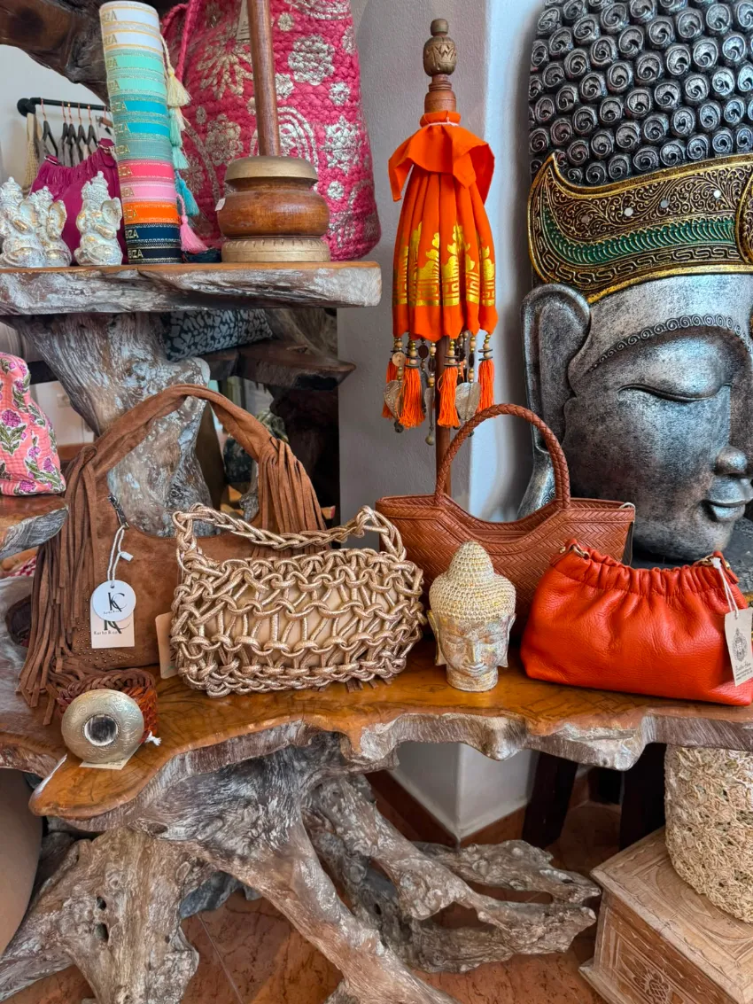 Exhibición decorativa de bolsos tejidos, pequeñas figuritas y una estatua de una cabeza de Buda sobre estantes rústicos de madera.