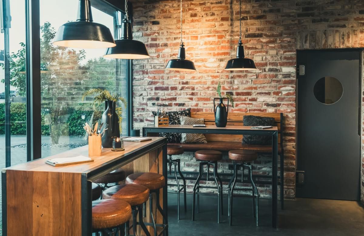 Interior bar with brick wall, wooden counter, black stools, pendant lights, and glass window.
