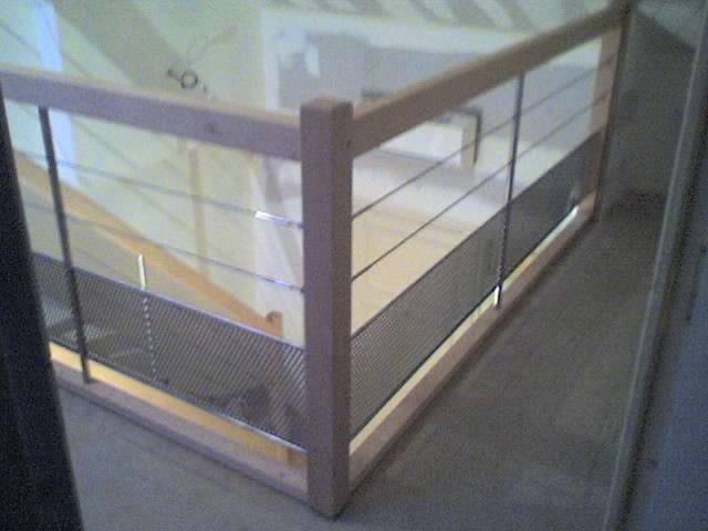 Rampe d'escalier en bois avec fils métalliques horizontaux, adjacente à un panneau en treillis métallique.